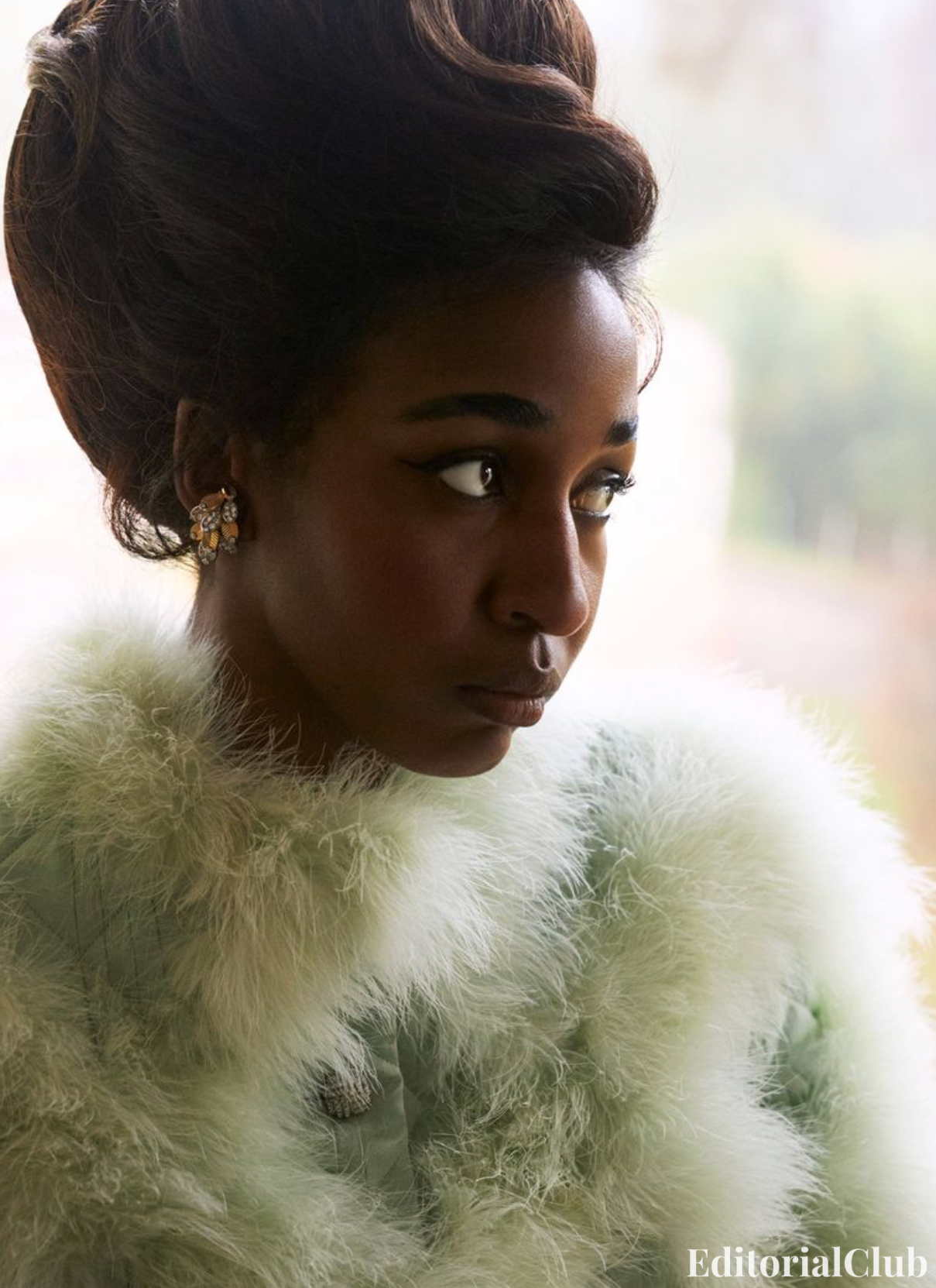 Ayo Edebiri in a mint green feathered coat with vintage-inspired updo, gazing out a window in soft natural light.