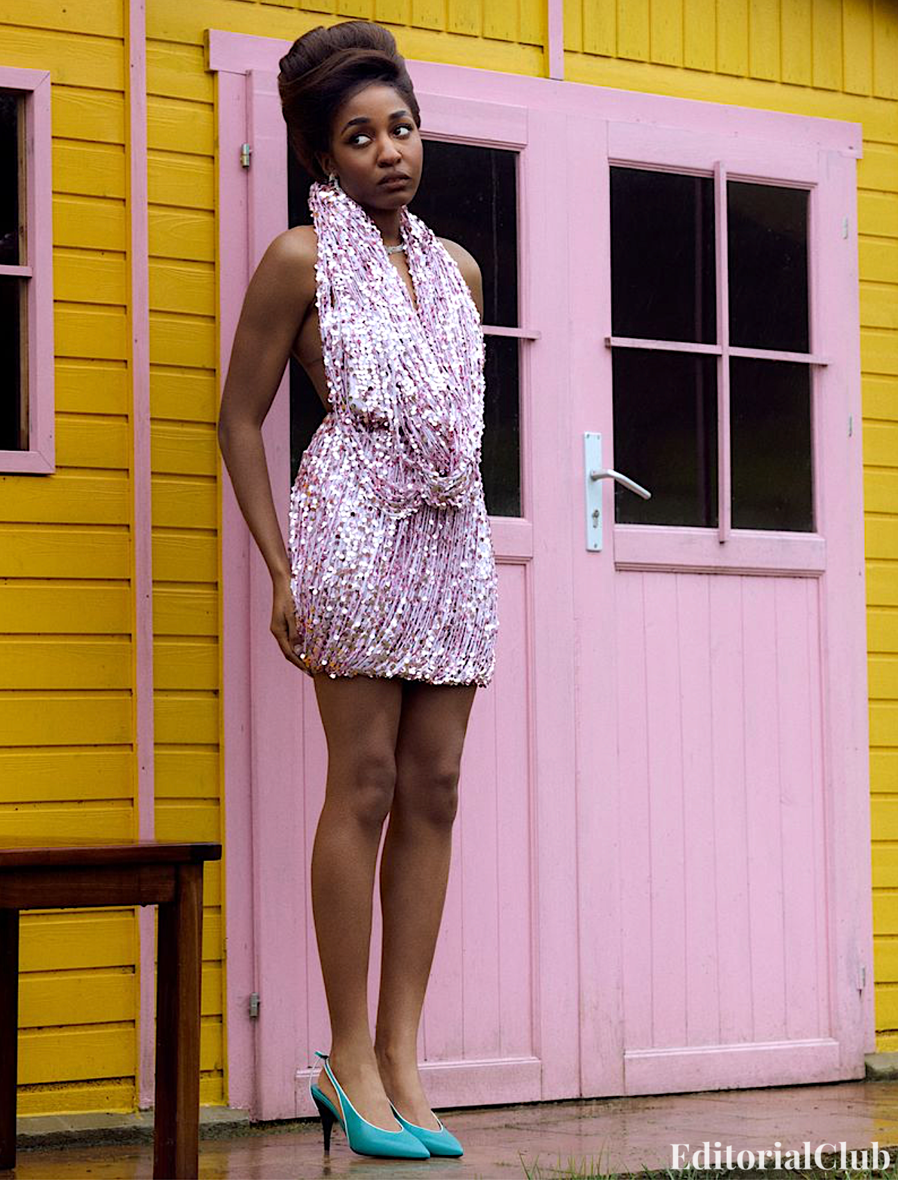 Ayo Edebiri stands in front of a pastel pink and yellow house in a shimmering pink mini dress and turquoise heels.