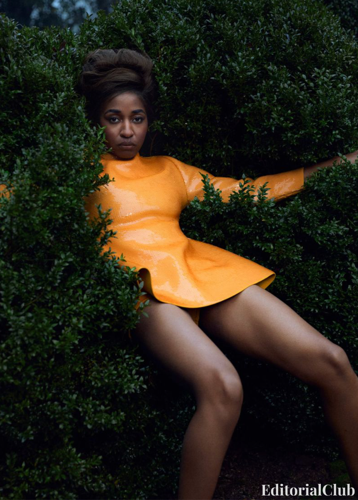 Ayo Edebiri poses confidently in a sculptural orange latex dress, framed by lush green hedges in a bold outdoor editorial shot.