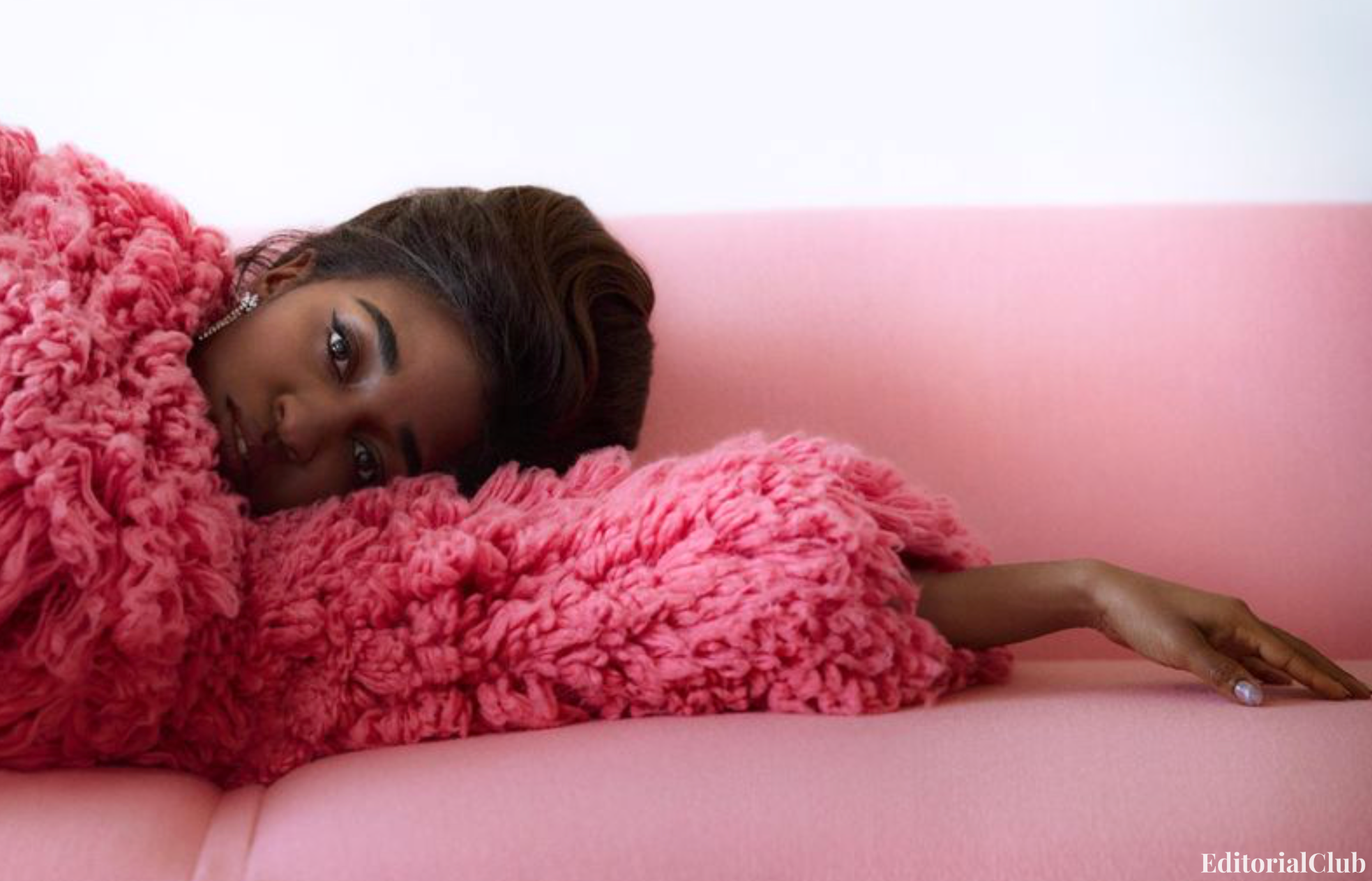 Ayo Edebiri reclines on a pink couch wrapped in a vivid pink fur coat, creating a soft and intimate fashion moment.