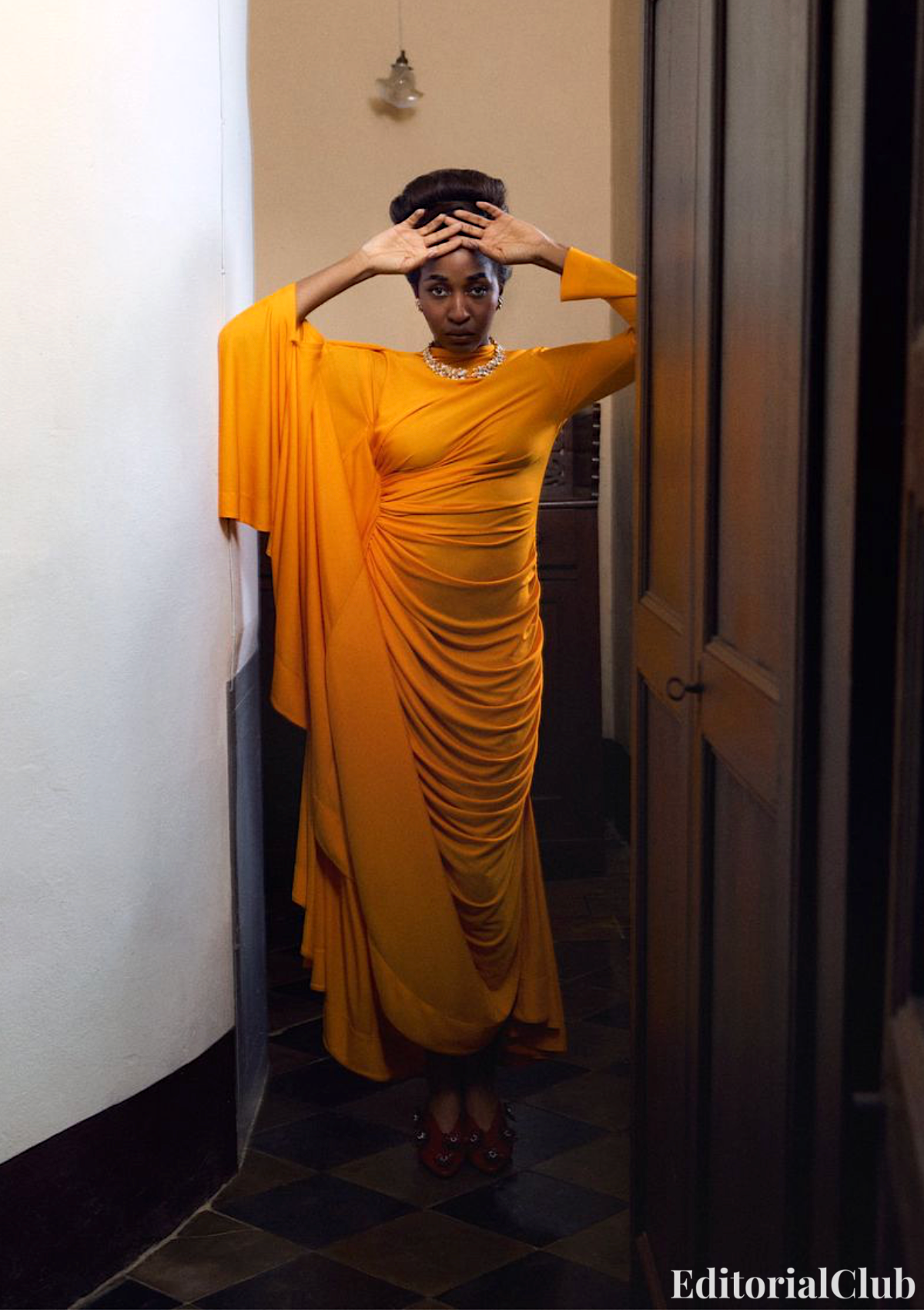 Ayo Edebiri in a draped orange gown with ruched details, striking a regal pose in a narrow vintage hallway.
