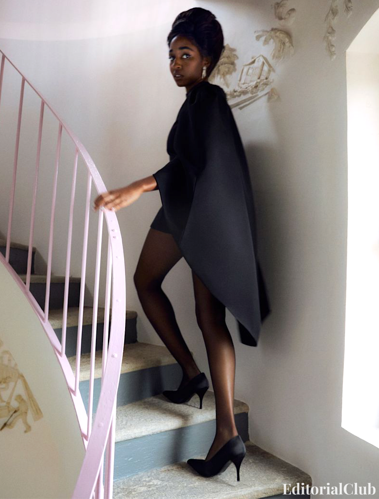 Ayo Edebiri walks up a spiral staircase in sheer black tights and a structured black cape dress, exuding classic elegance.