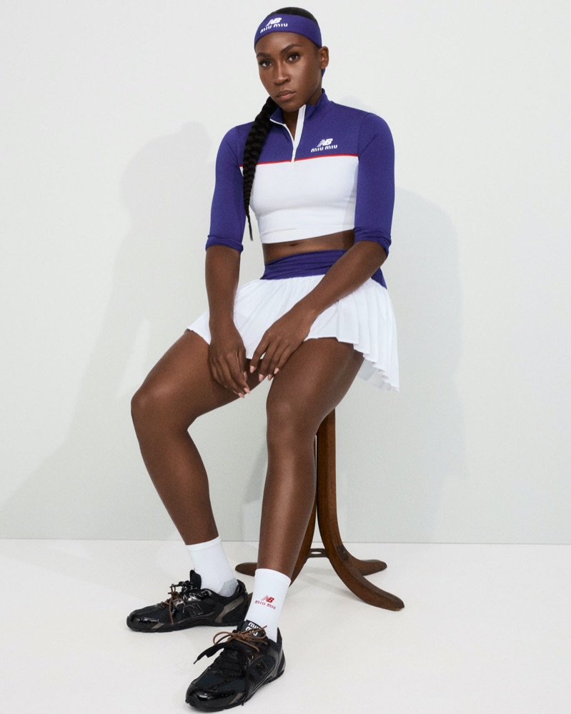 Coco Gauff seated on a wooden stool in a white and purple Miu Miu x New Balance crop top and pleated skirt with black sneakers