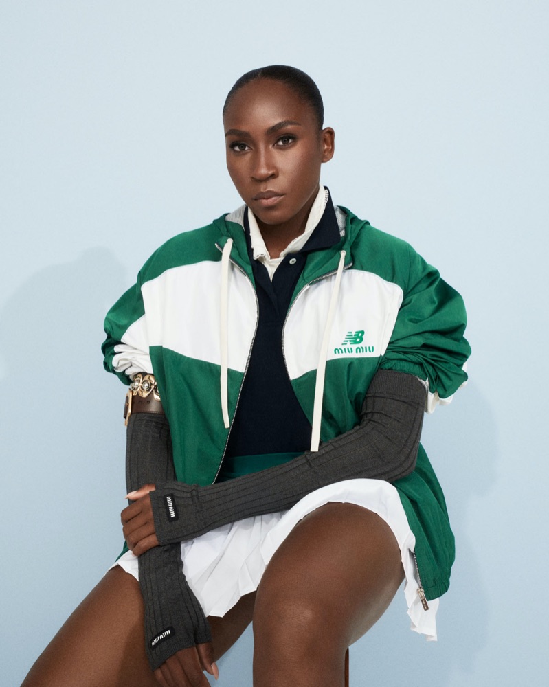 Coco Gauff seated in a white pleated skirt and green Miu Miu x New Balance jacket with layered styling and ribbed gloves