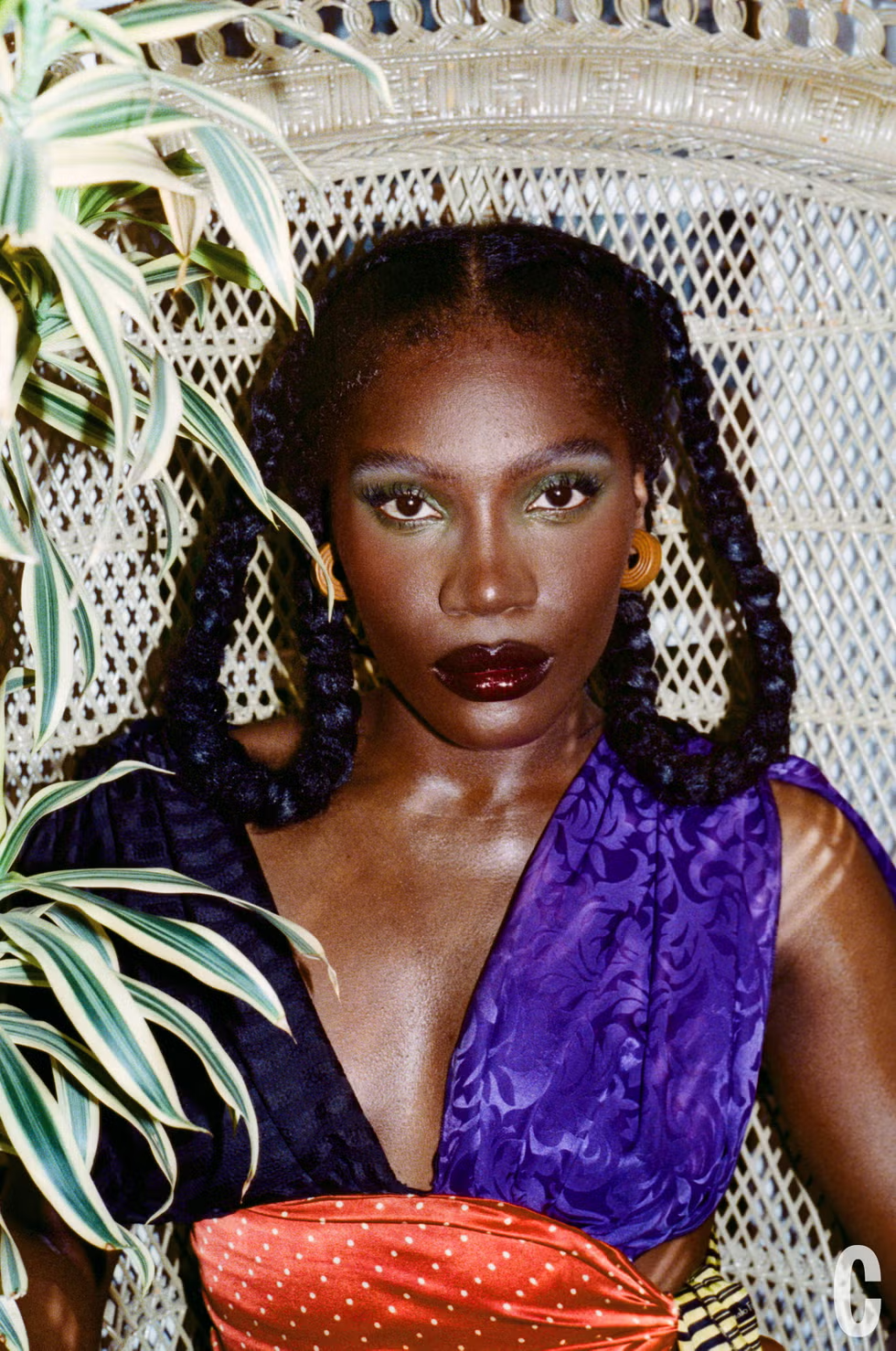 Close-up of Doechii with bold green eyeshadow, glossy lips, and braided hair, wearing a purple and red patterned top against a rattan backdrop, Cosmopolitan US April 2025.