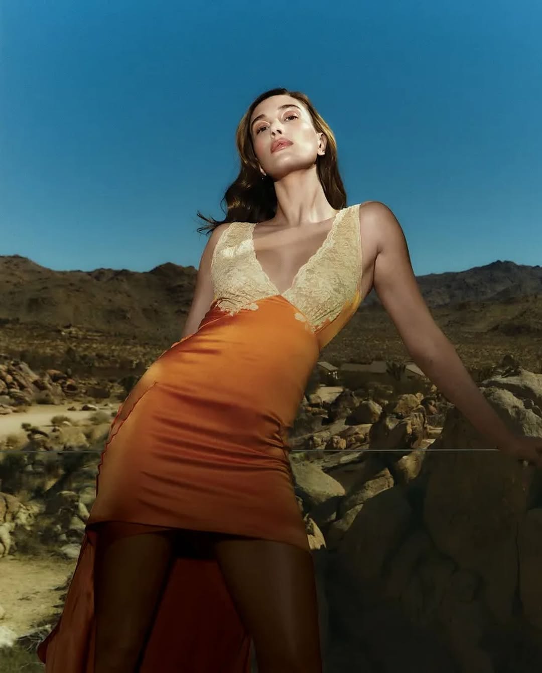 Hailey Bieber in deep orange satin dress with lace bodice, posing confidently against a desert landscape under a clear blue sky