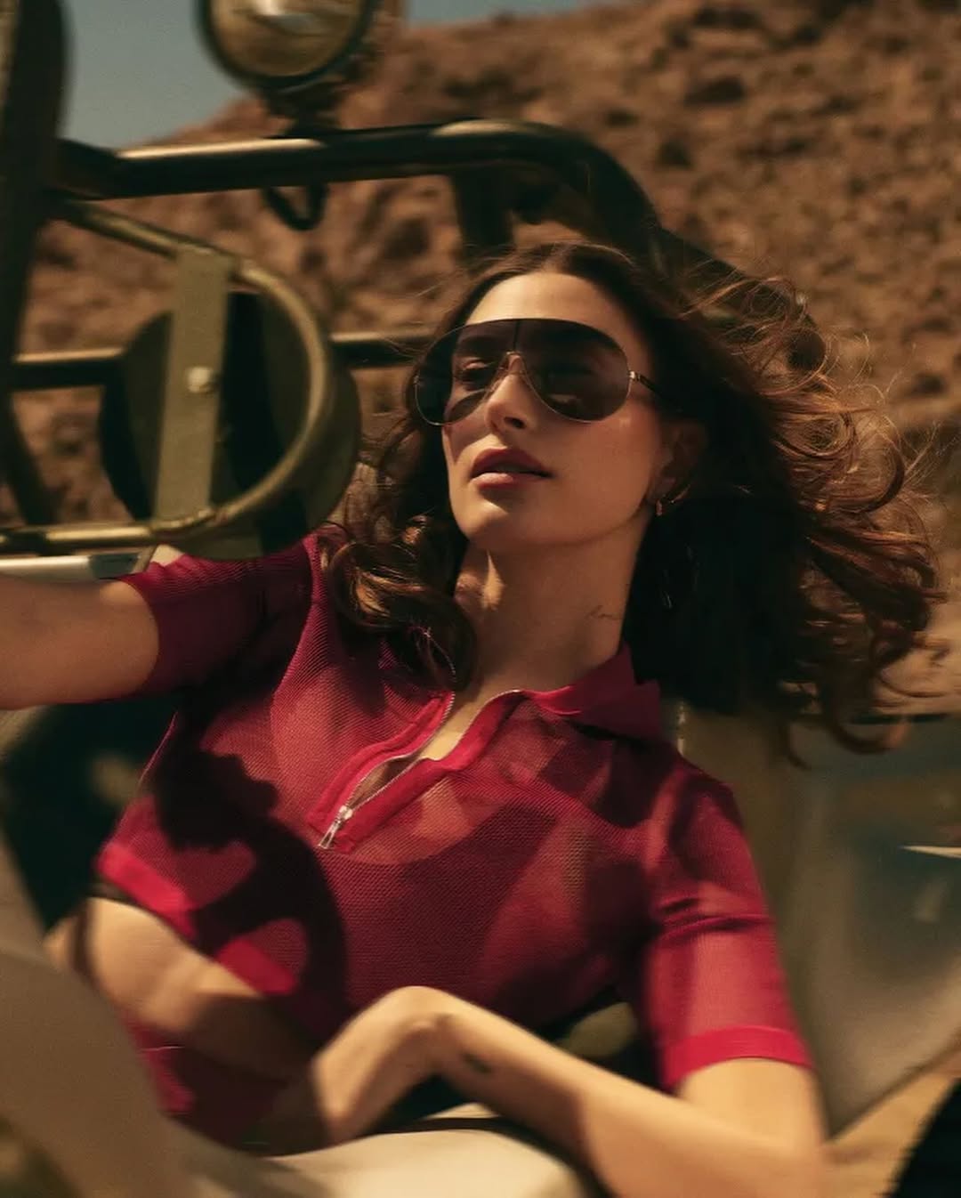 Hailey Bieber driving a vintage vehicle in the desert, wearing a sheer red zip-up top and oversized sunglasses, wind in her hair
