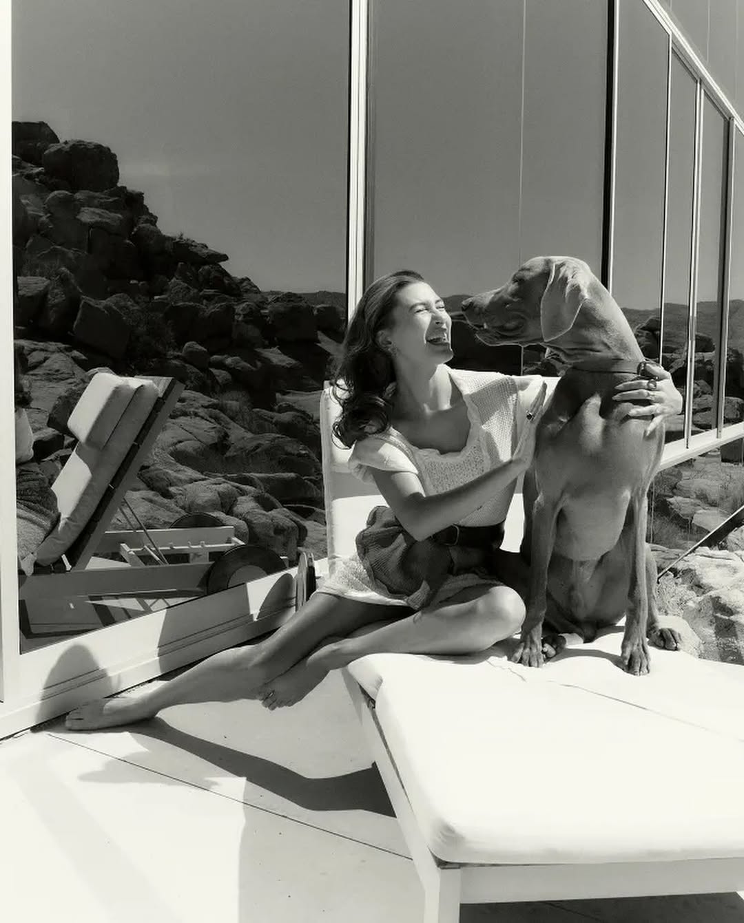 Black and white image of Hailey Bieber laughing while lounging with a large dog on a white outdoor chaise in front of a glass house