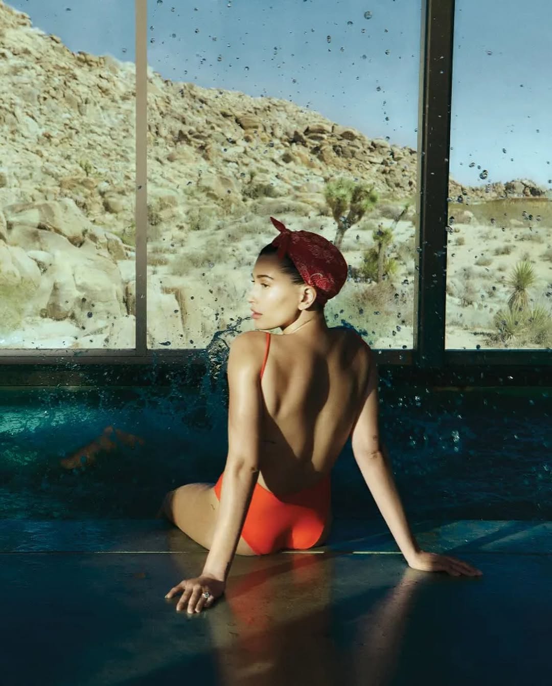 Hailey Bieber sitting poolside in a red one-piece swimsuit with a red scarf in her hair, looking out at the rocky desert through a glass window