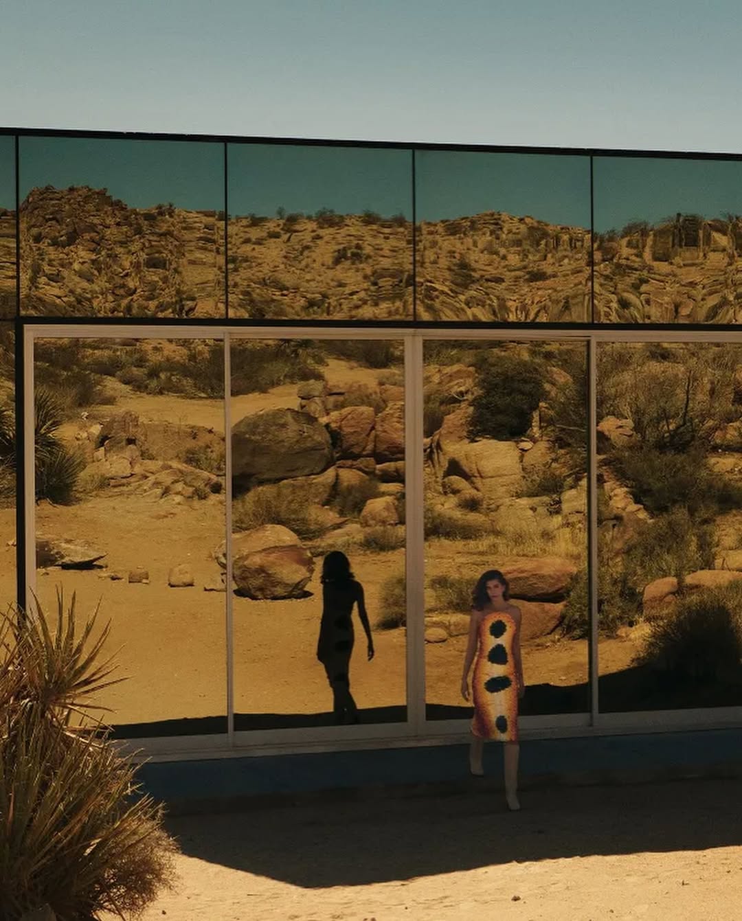 Hailey Bieber in a yellow dress with black spots walking in front of a glass building reflecting the desert, casting a strong silhouette