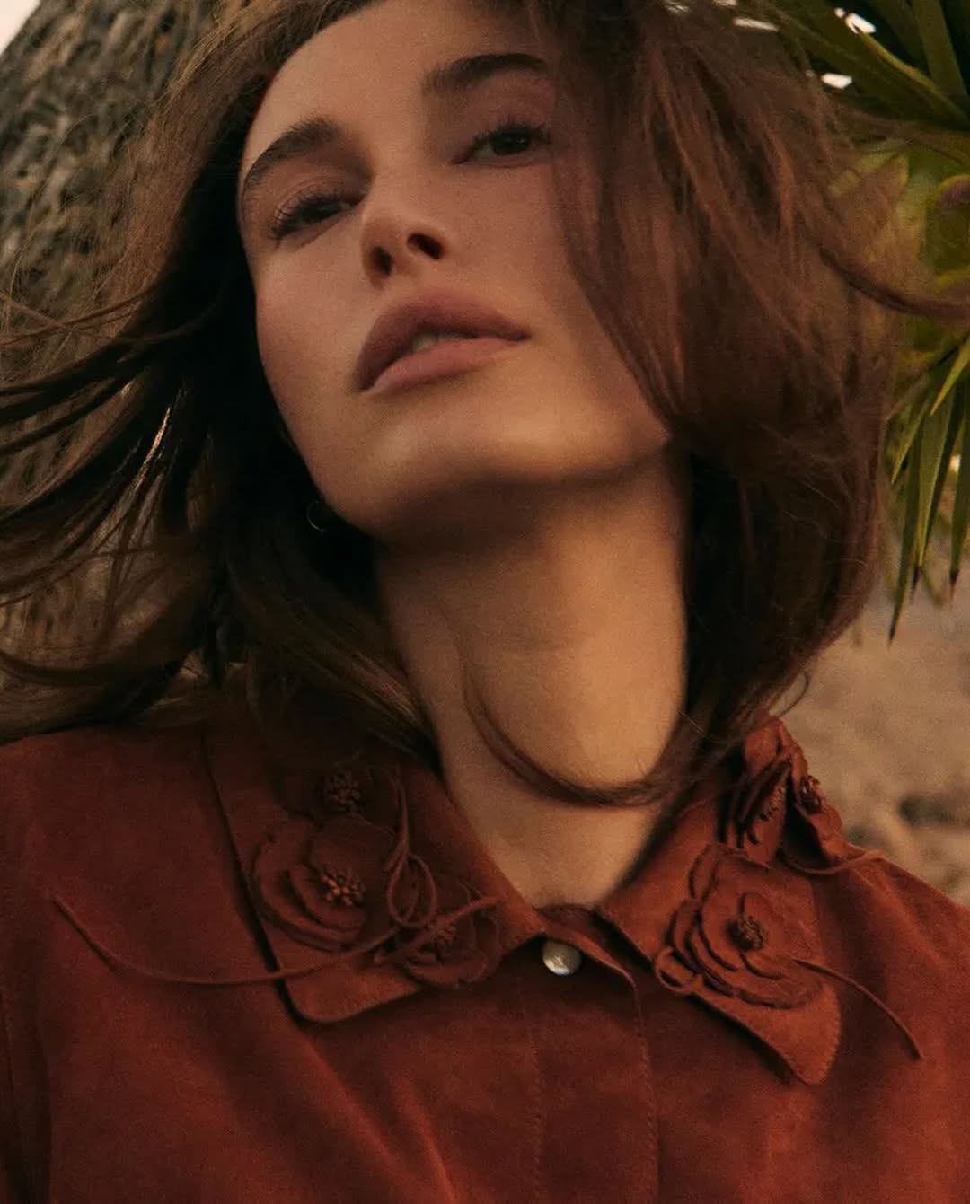 Close-up portrait of Hailey Bieber in a suede collared dress with floral embroidery, gazing upward in soft desert light