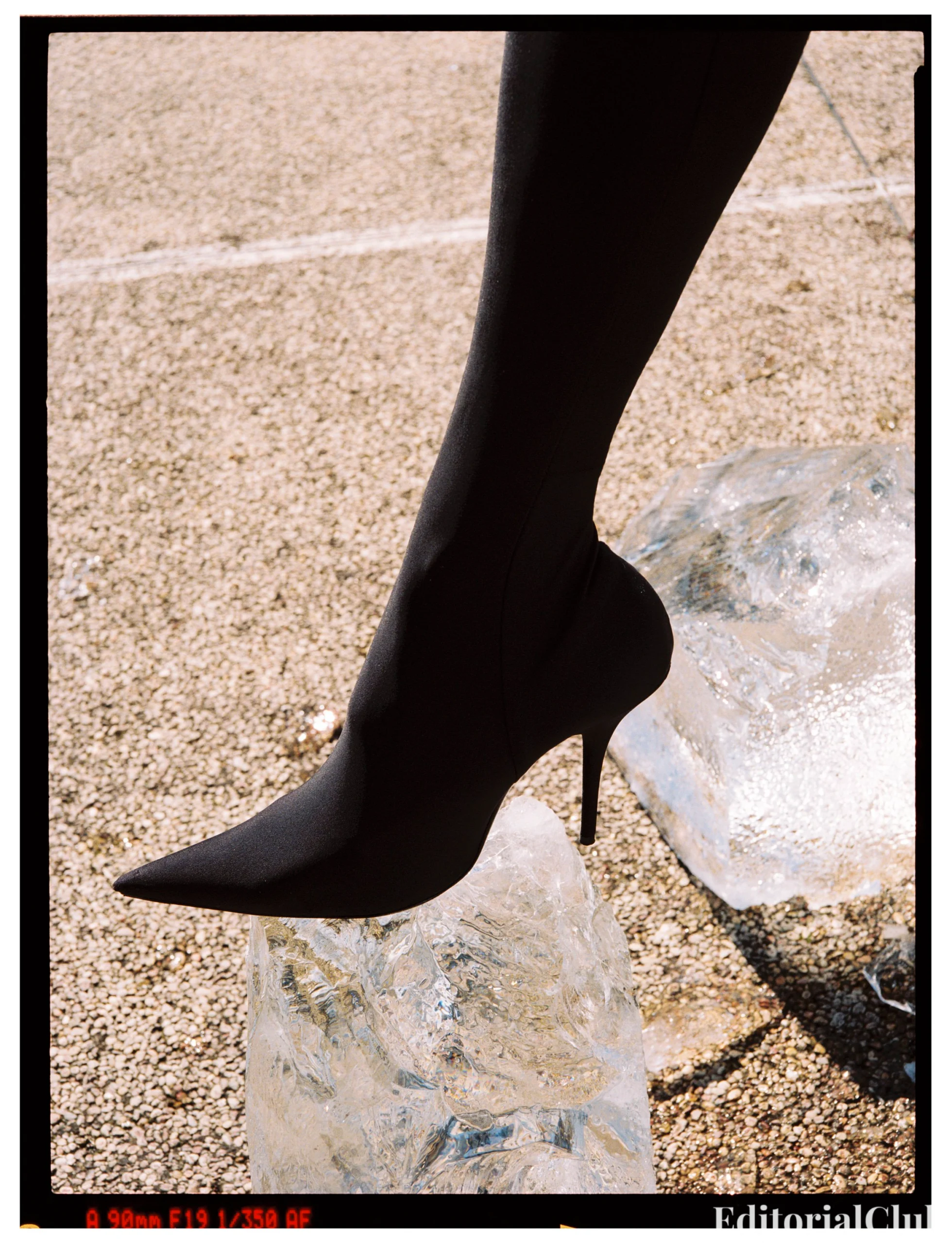 Close-up of Isabeli Fontana's foot in a pointed black stiletto boot balanced on a melting ice block outdoors.