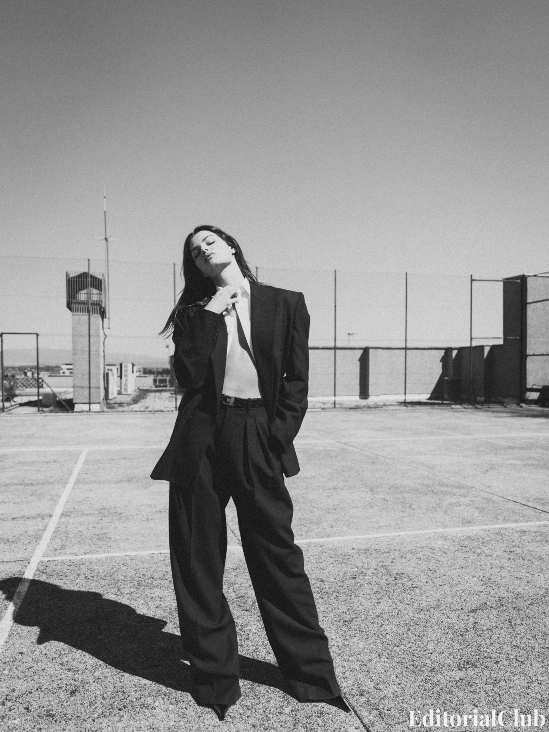 Isabeli Fontana wearing an oversized black suit on an urban rooftop, eyes closed and chin tilted back under the sun.