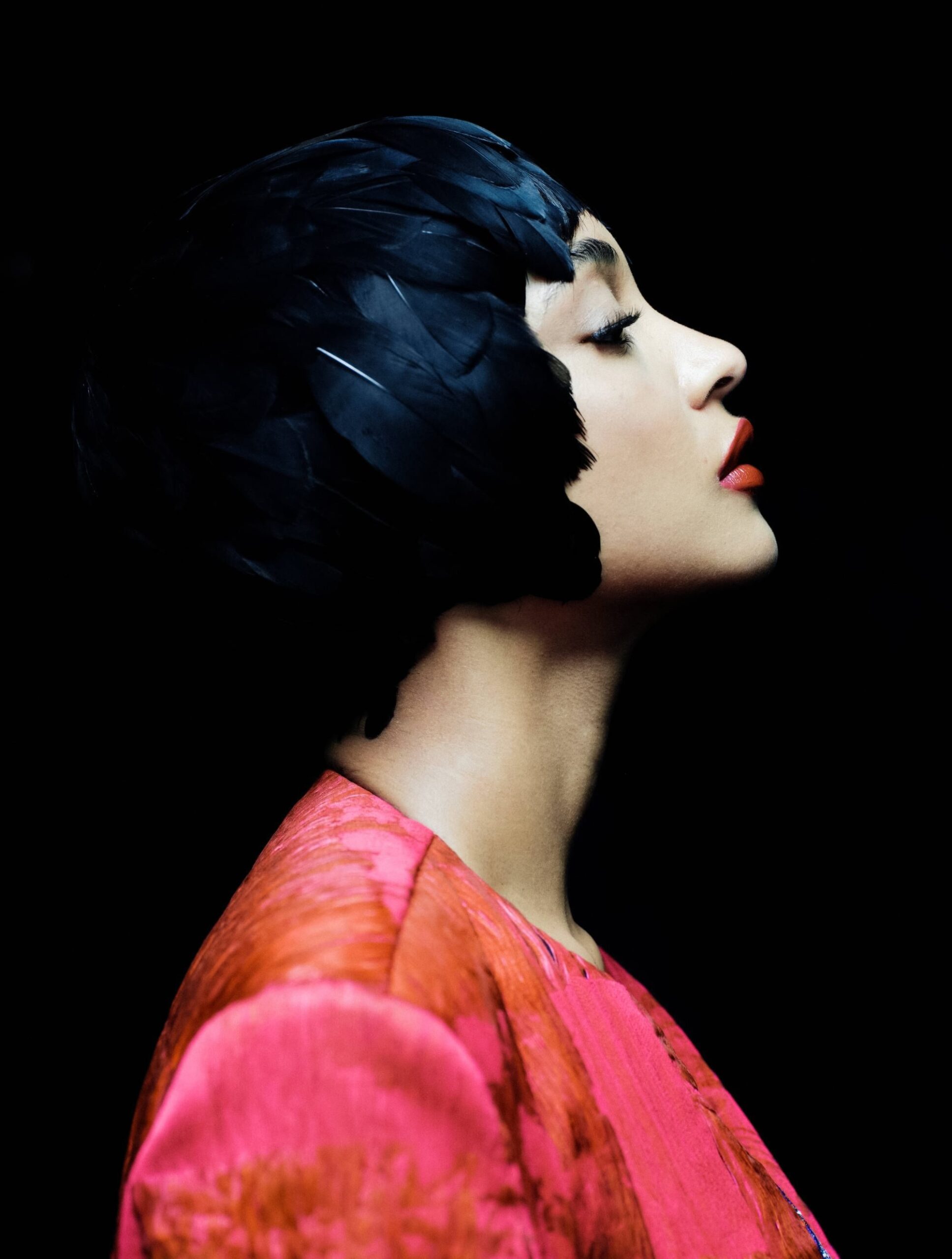 Side profile of Jourdan Dunn wearing a feathered black headpiece and red textured outfit in dark lighting