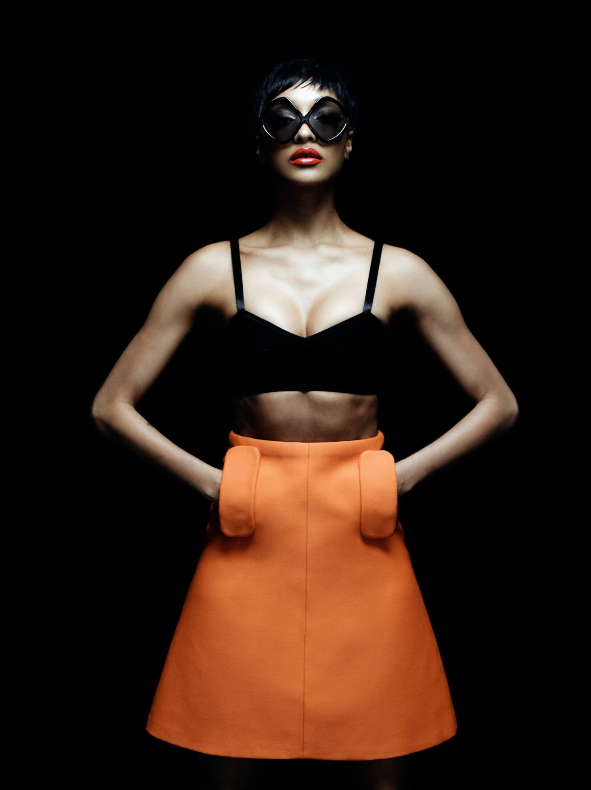 Jourdan Dunn posing against a black background in oversized sunglasses, black crop top, and orange skirt for Harper’s Bazaar España 2025.