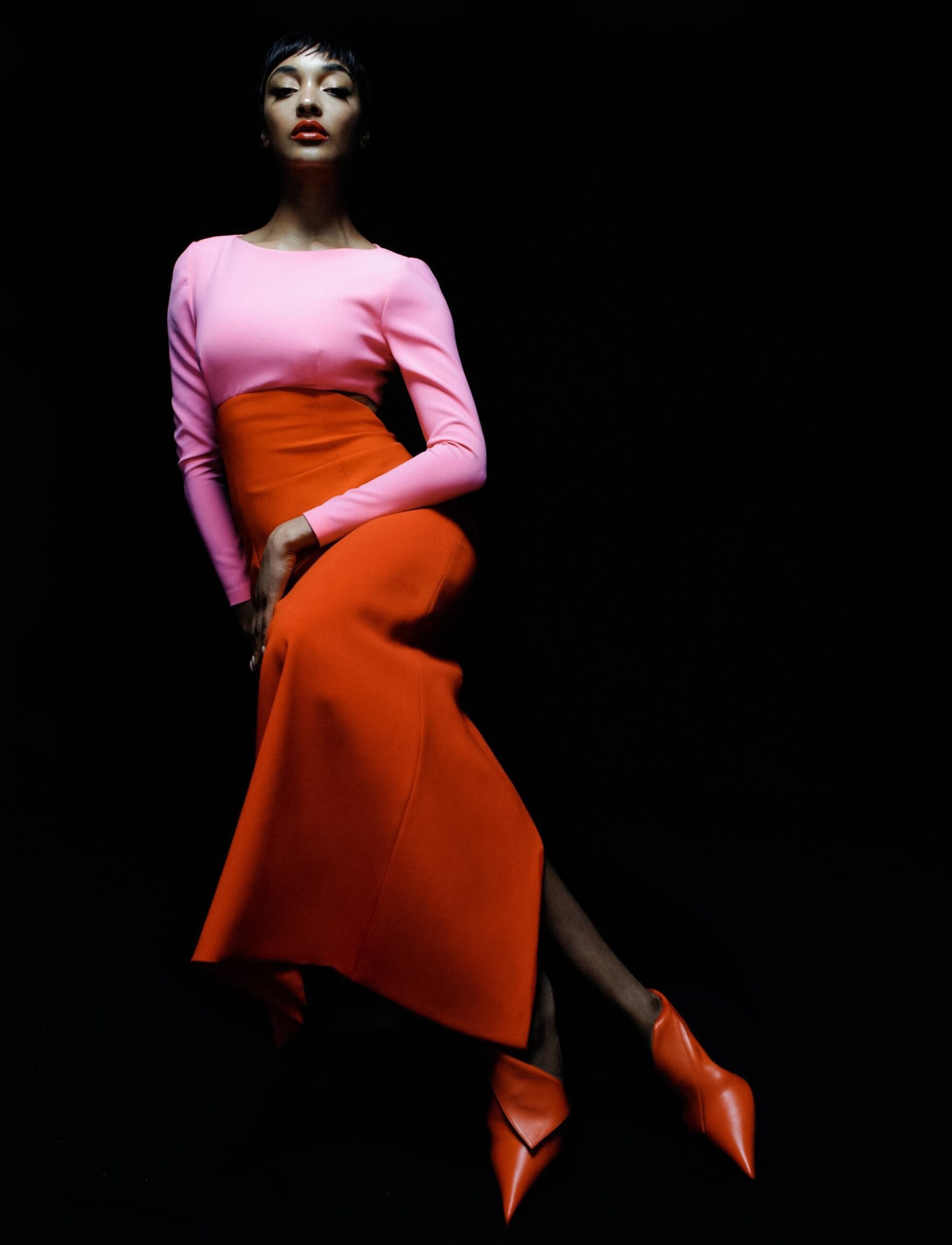 Jourdan Dunn wearing a color-blocked pink and red gown with pointed red heels, photographed on a black backdrop for Harper’s Bazaar España.