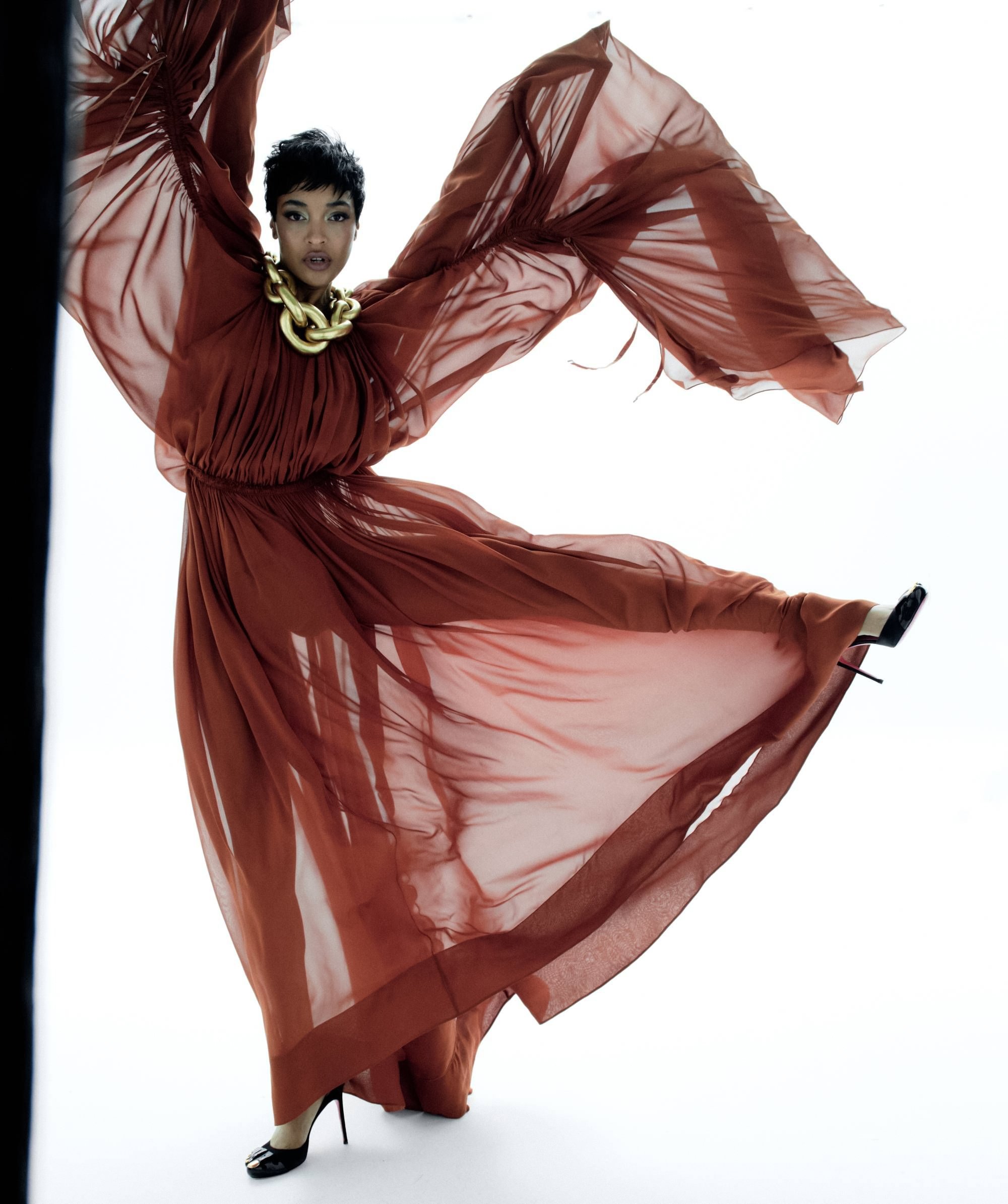 Jourdan Dunn in a flowing rust-red gown with exaggerated sleeves and chunky gold necklace, captured mid-motion for Harper’s Bazaar España May 2025.