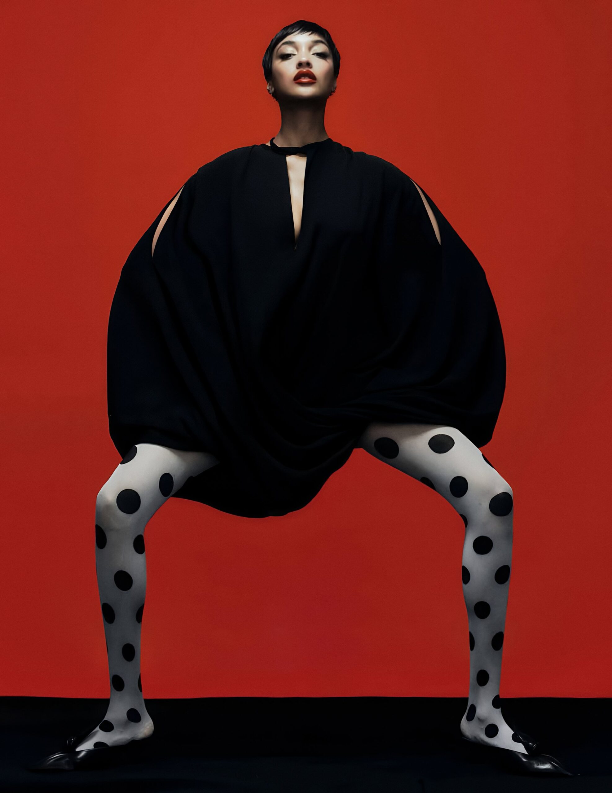 Jourdan Dunn in a sculptural black dress and polka-dot tights against a vivid red background