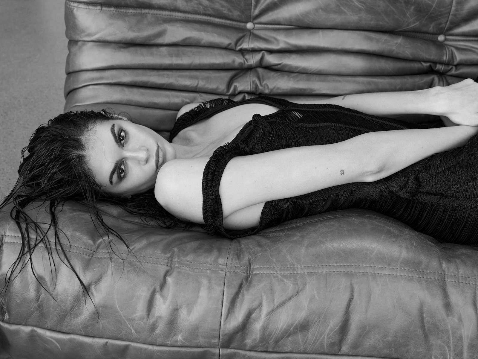 Kaia Gerber lounges in a low-cut black dress on a leather sofa in a moody black and white shot