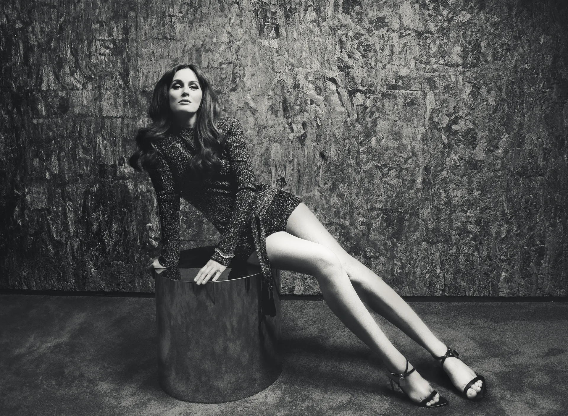 Leighton Meester reclines on a chrome cylinder stool in a shimmering mini dress, shot against a dark textured wall in a high-fashion black-and-white photo.