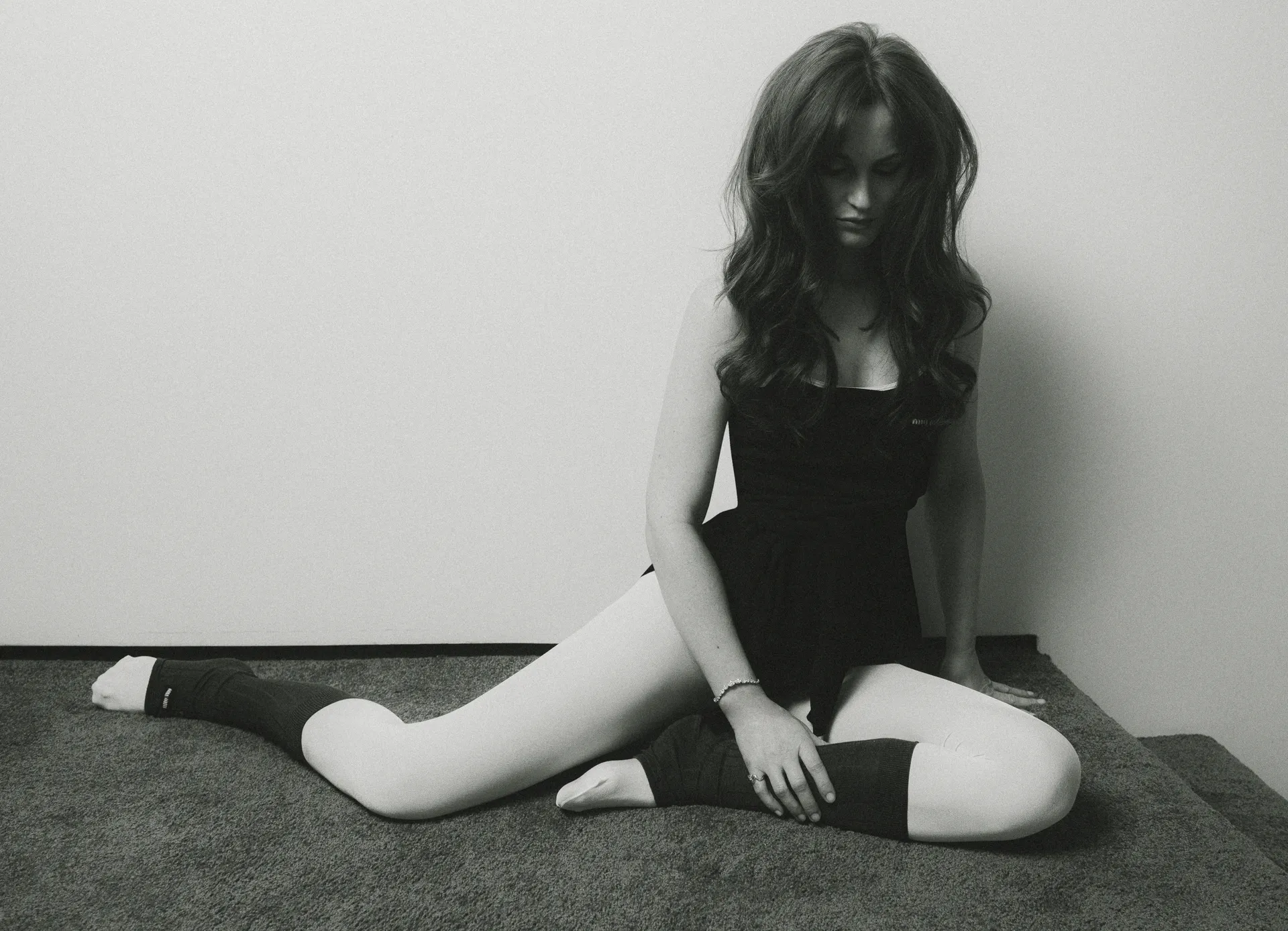 Leighton Meester sits on the floor in a black bodysuit and white tights with leg warmers, gazing down in a moody monochrome editorial image.
