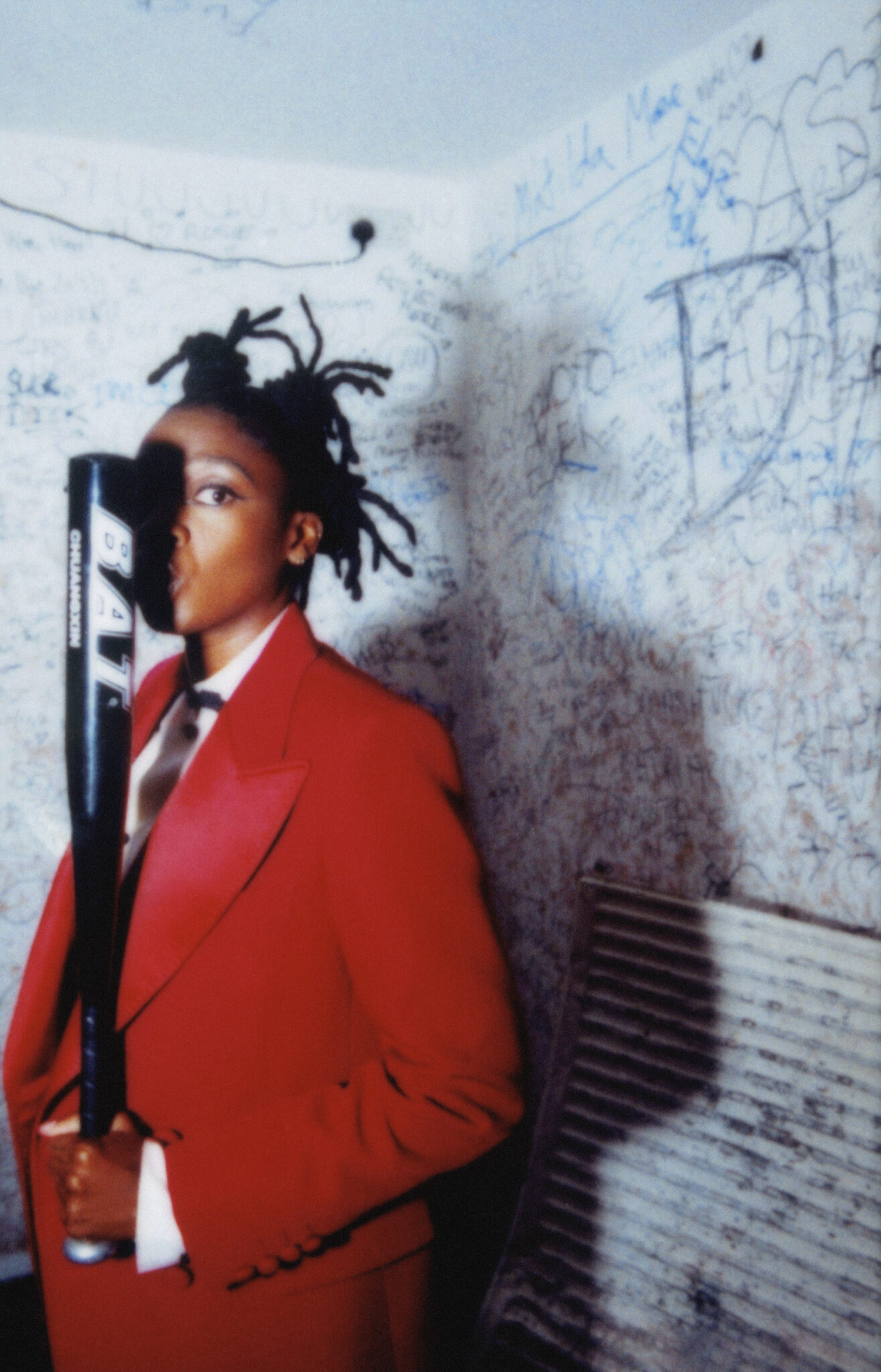 Portrait of Little Simz in a bold red suit holding a black baseball bat, standing in a gritty graffiti-covered corner, Interview Magazine Spring 2025.