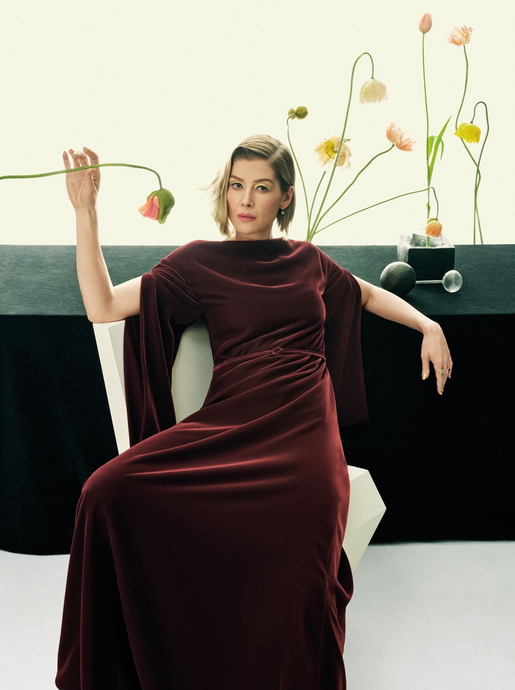 Rosamund Pike in a deep red velvet gown seated beside pastel poppies in a modern editorial setting