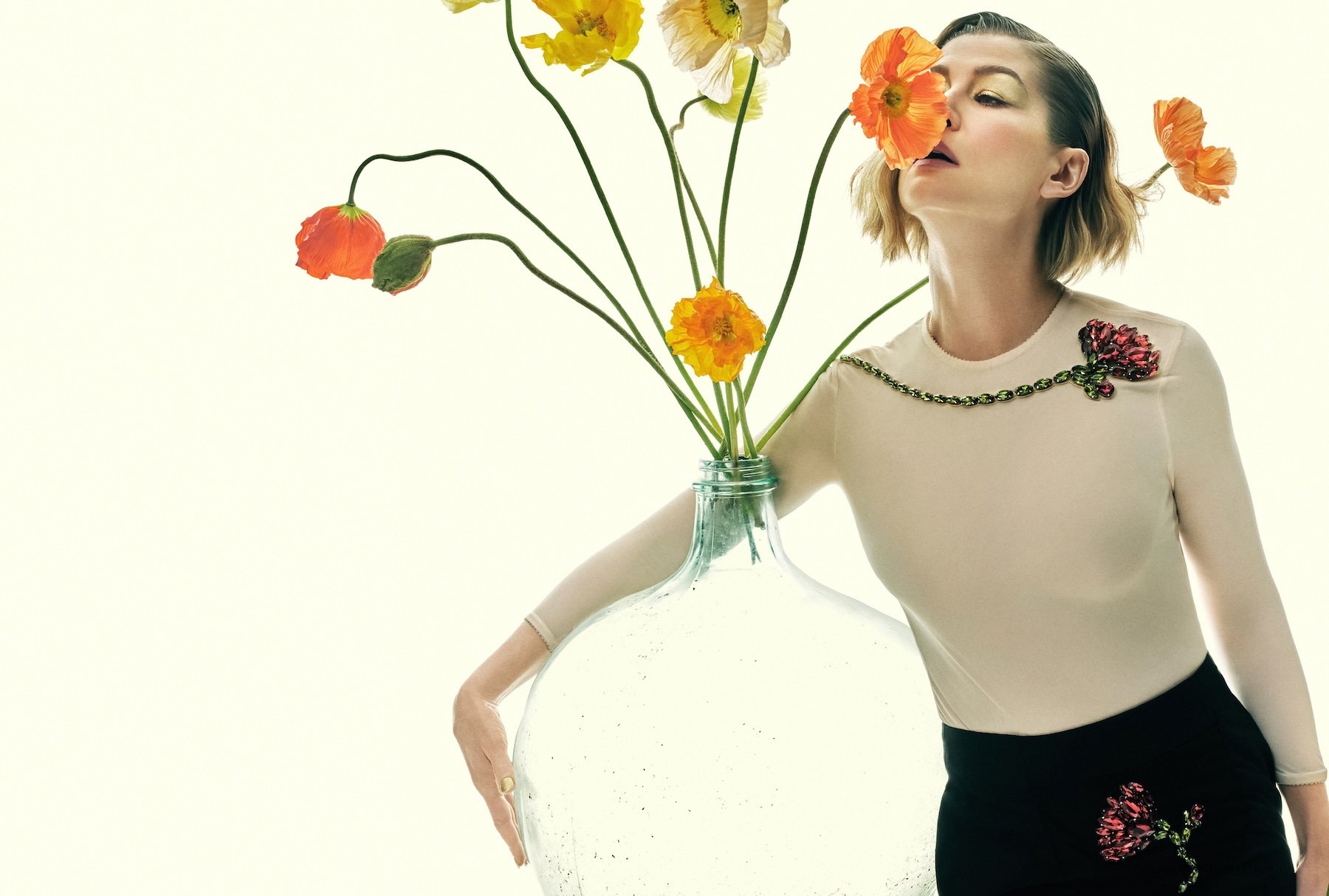 Rosamund Pike leaning against a table with pastel orange poppies while wearing a sheer nude top with floral embellishments