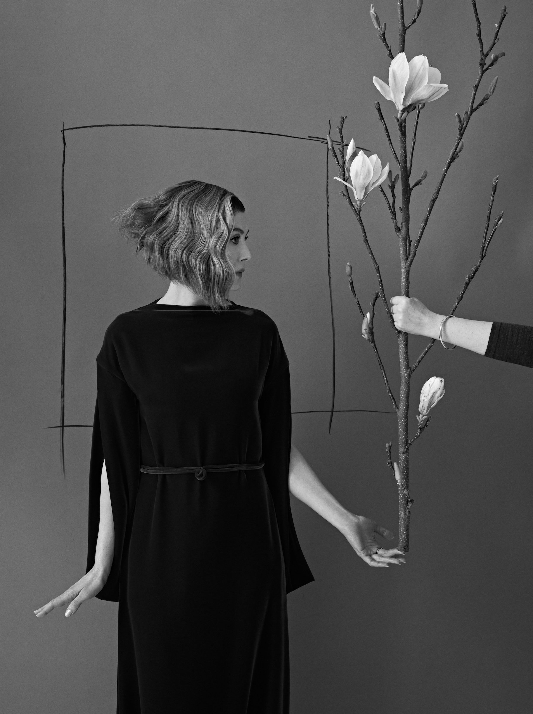 Rosamund Pike in a black gown standing beside a magnolia branch held by a stylist, set against a grey studio backdrop
