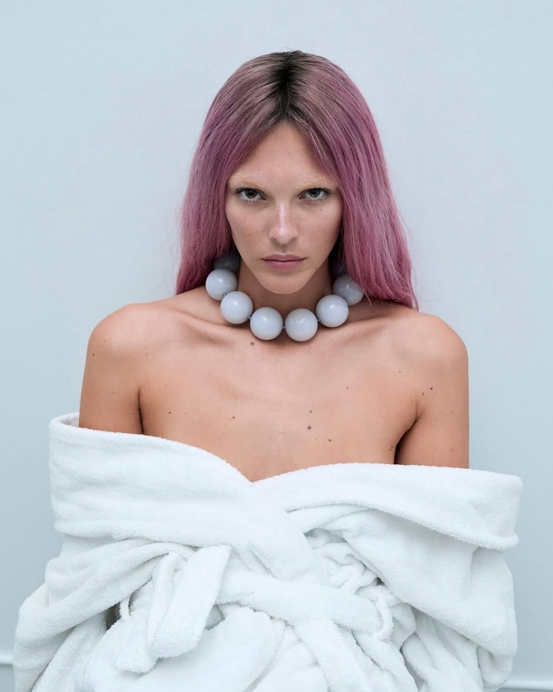 Vittoria Ceretti stares intensely wearing a white bathrobe and chunky gray necklace, framed against a soft blue background