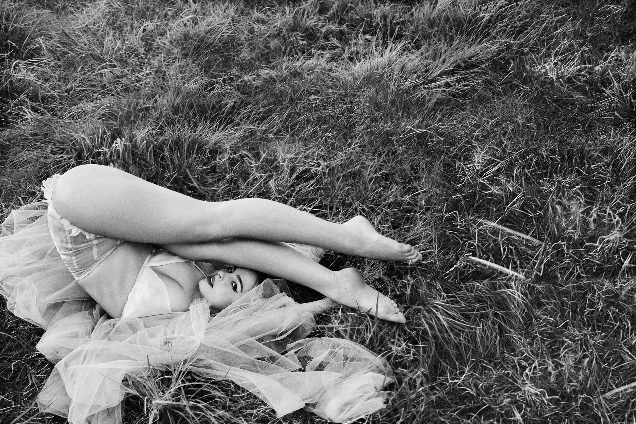 Black and white photo of Addison Rae playfully lying in the grass with her legs over her head, wearing sheer tulle and a white bra
