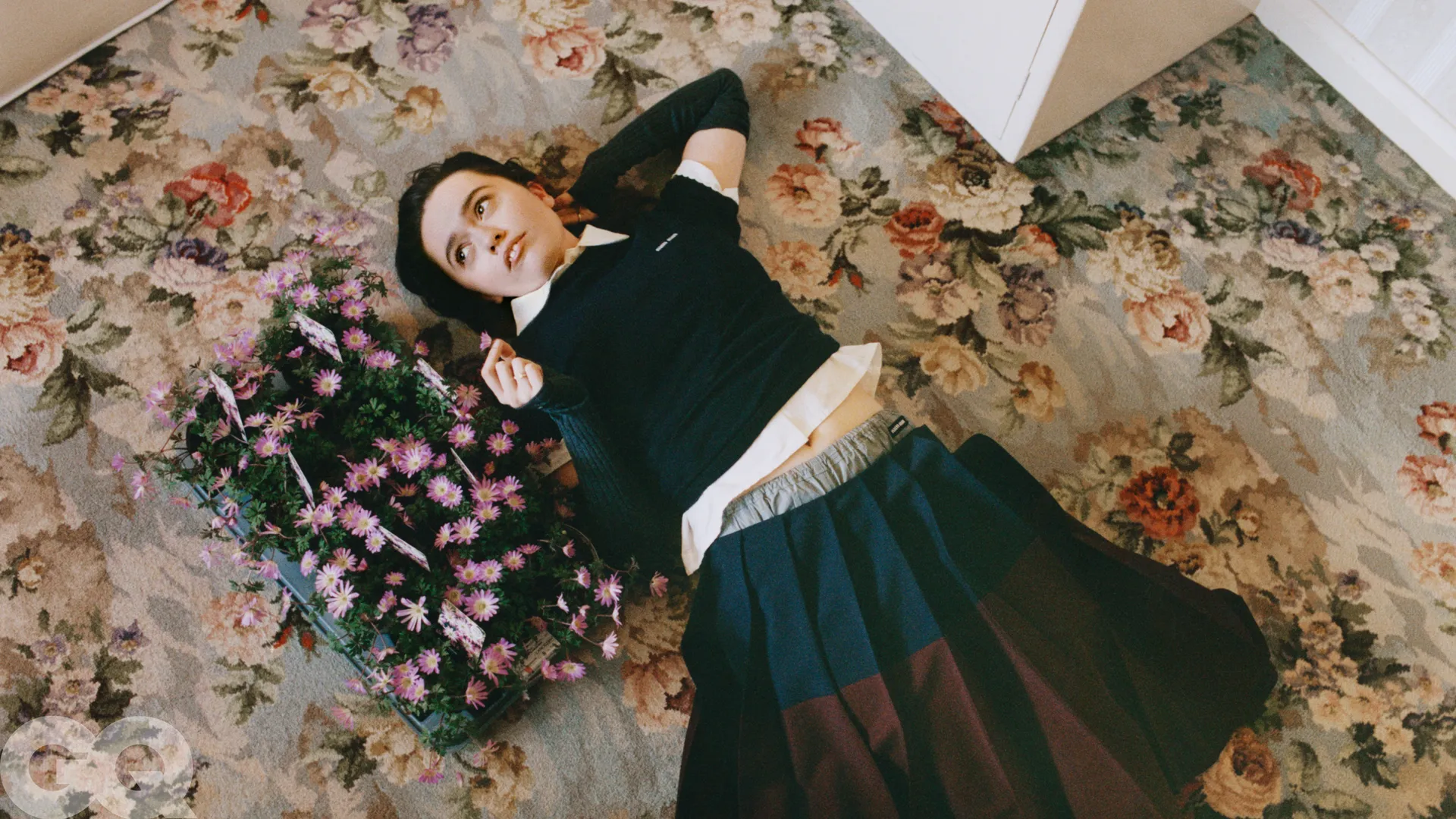Aimee Lou Wood lying on a vintage floral carpet next to a tray of purple daisies, wearing a navy and white layered outfit with a pleated skirt.