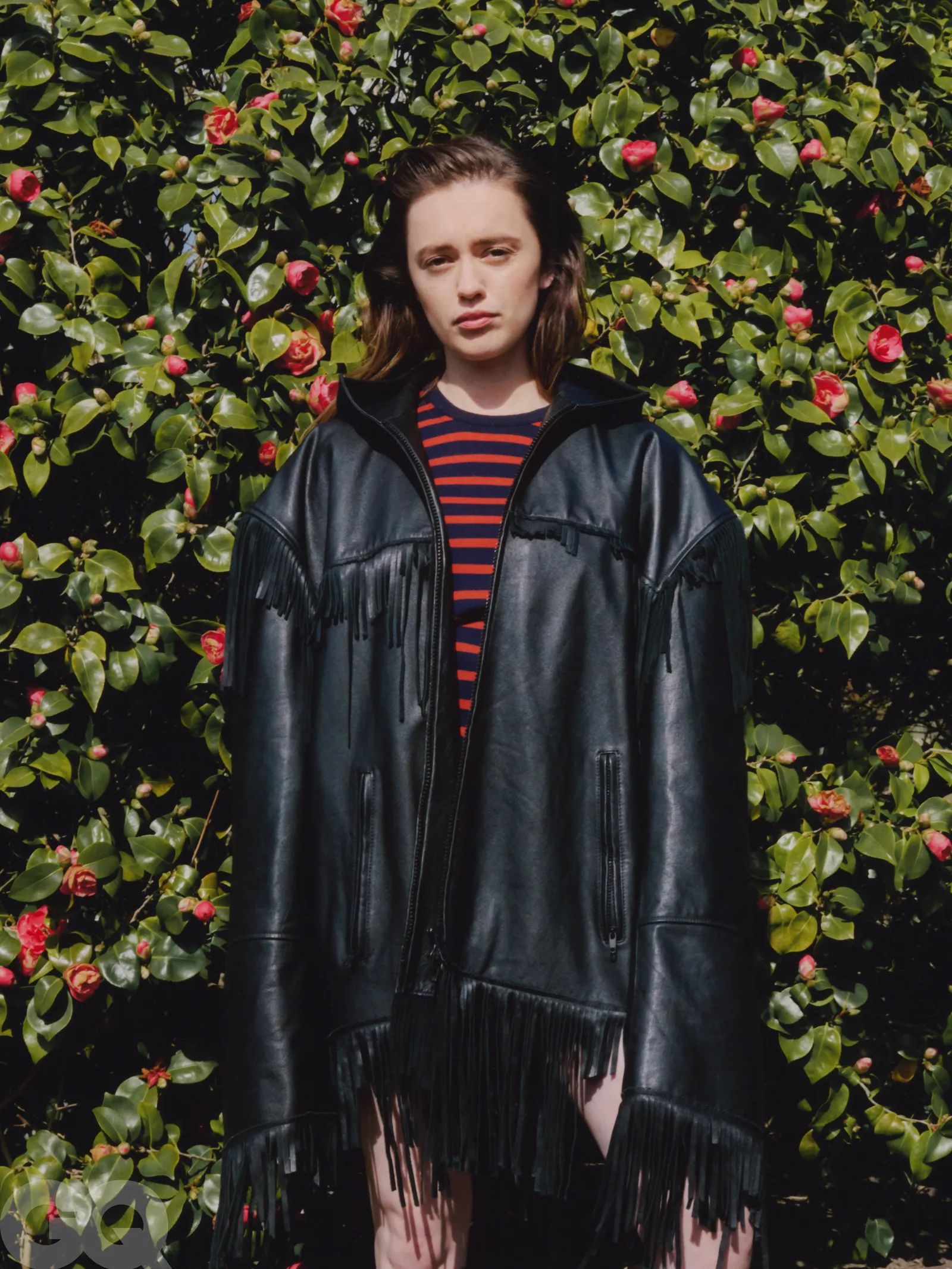 Aimee Lou Wood standing in front of a blooming camellia bush, wearing a black leather fringe coat over a striped top.
