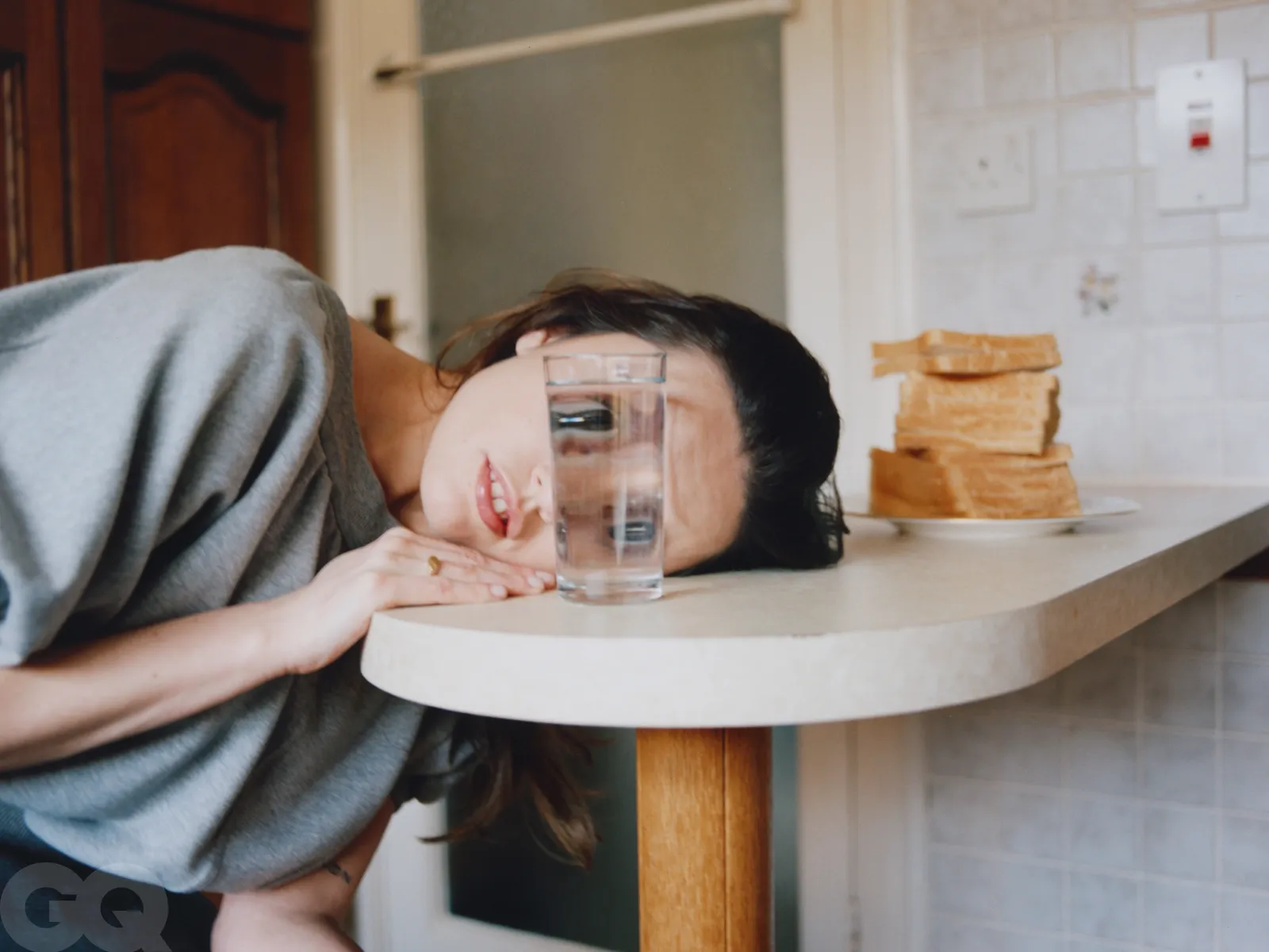 Aimee Lou Wood resting her face on a kitchen counter, peering through a glass of water with stacked toast slices in the background.
