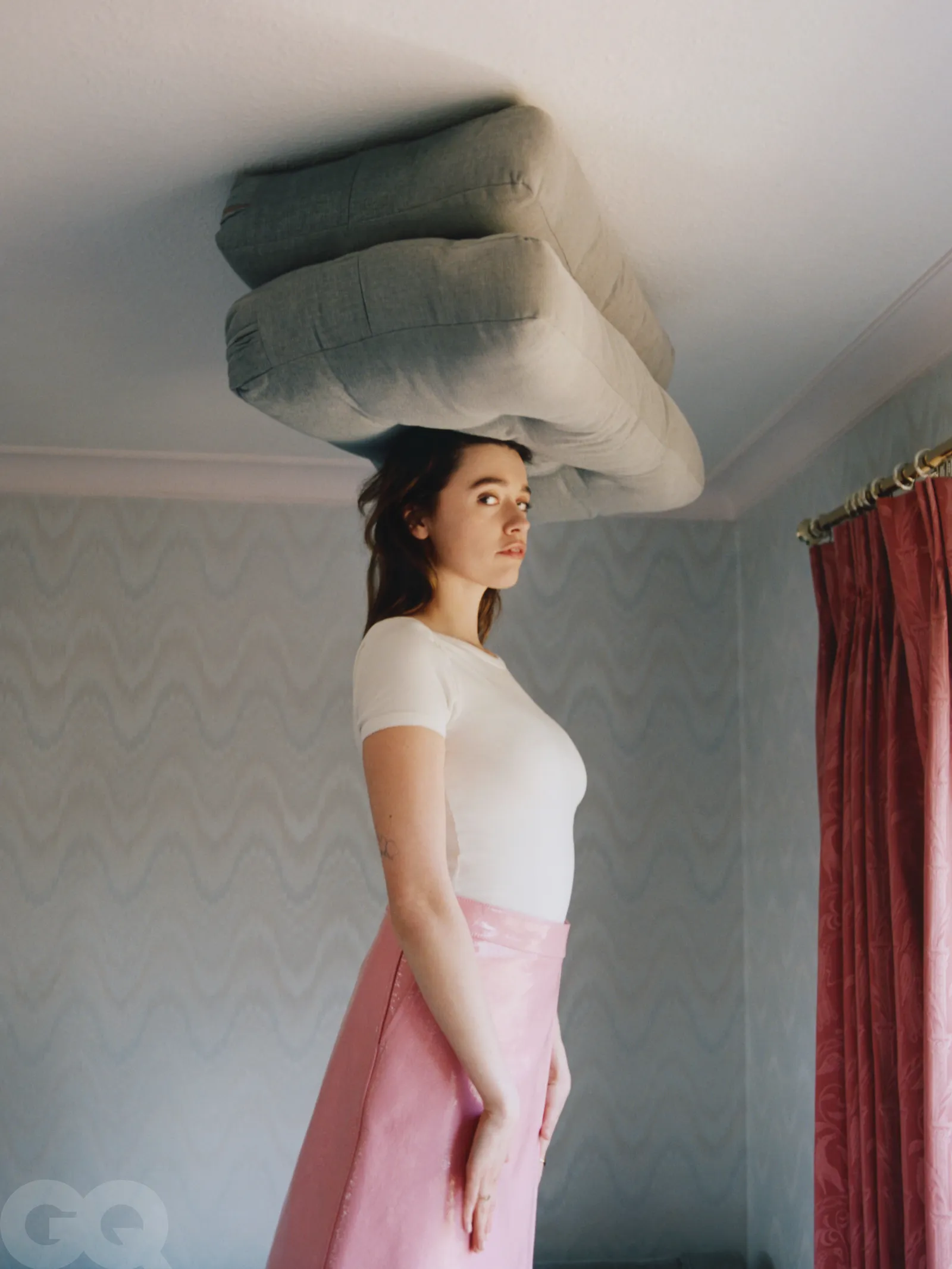 Aimee Lou Wood balancing gray cushions on her head while wearing a white top and pink skirt in a room with pastel wallpaper and curtains.