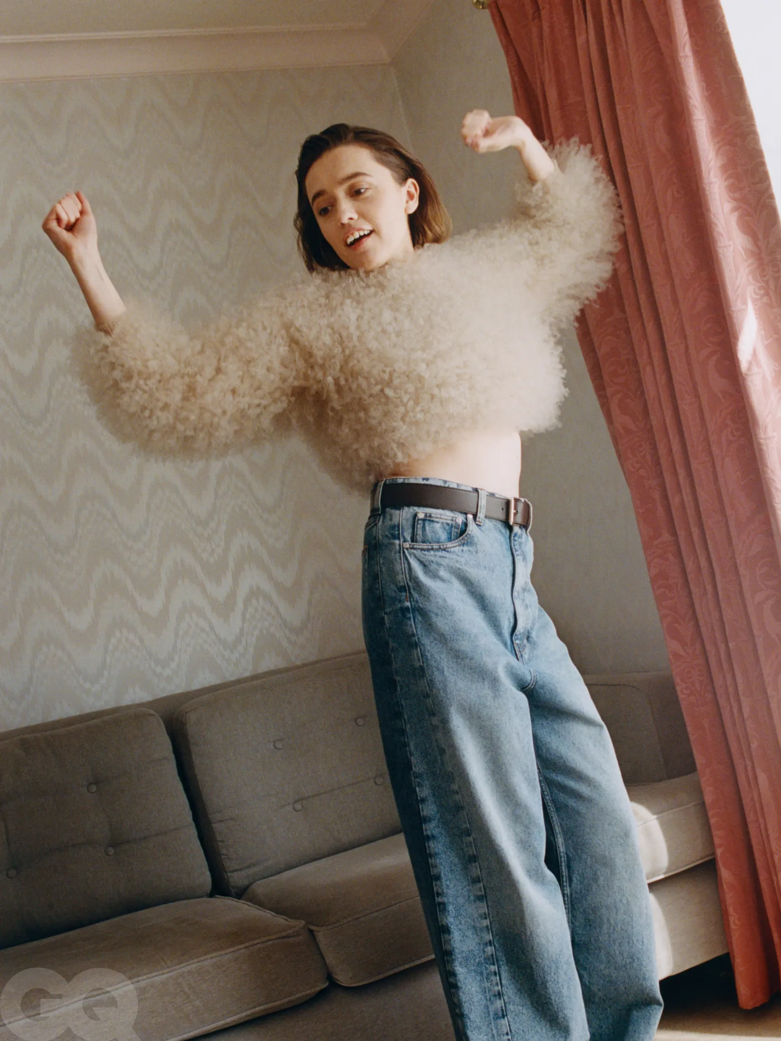 Aimee Lou Wood mid-movement in a beige cropped fuzzy sweater and wide-leg jeans, standing in a retro-styled living room.