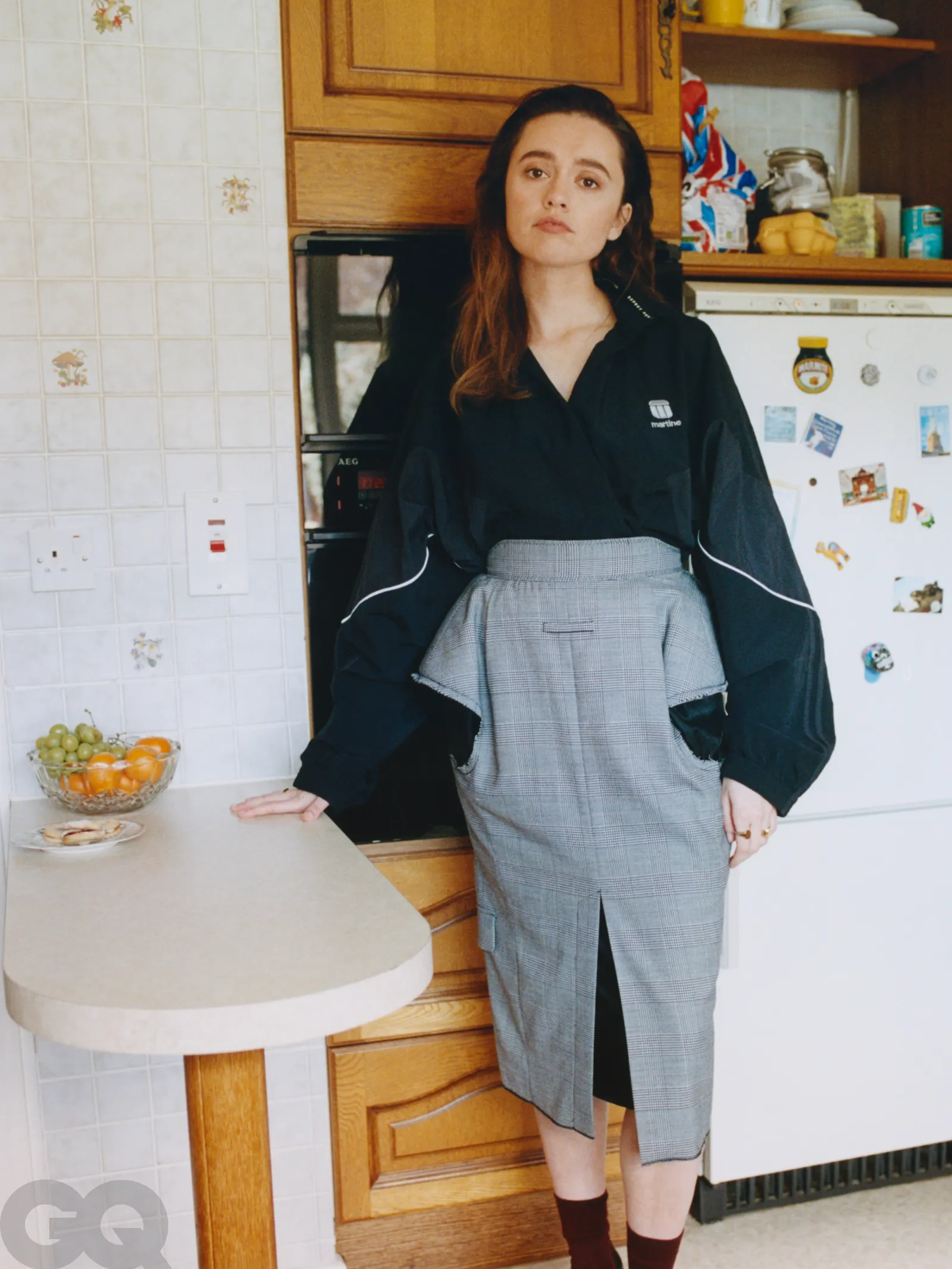 Aimee Lou Wood posing in a kitchen, wearing a structured gray pencil skirt and oversized black blouse, leaning on a vintage counter.