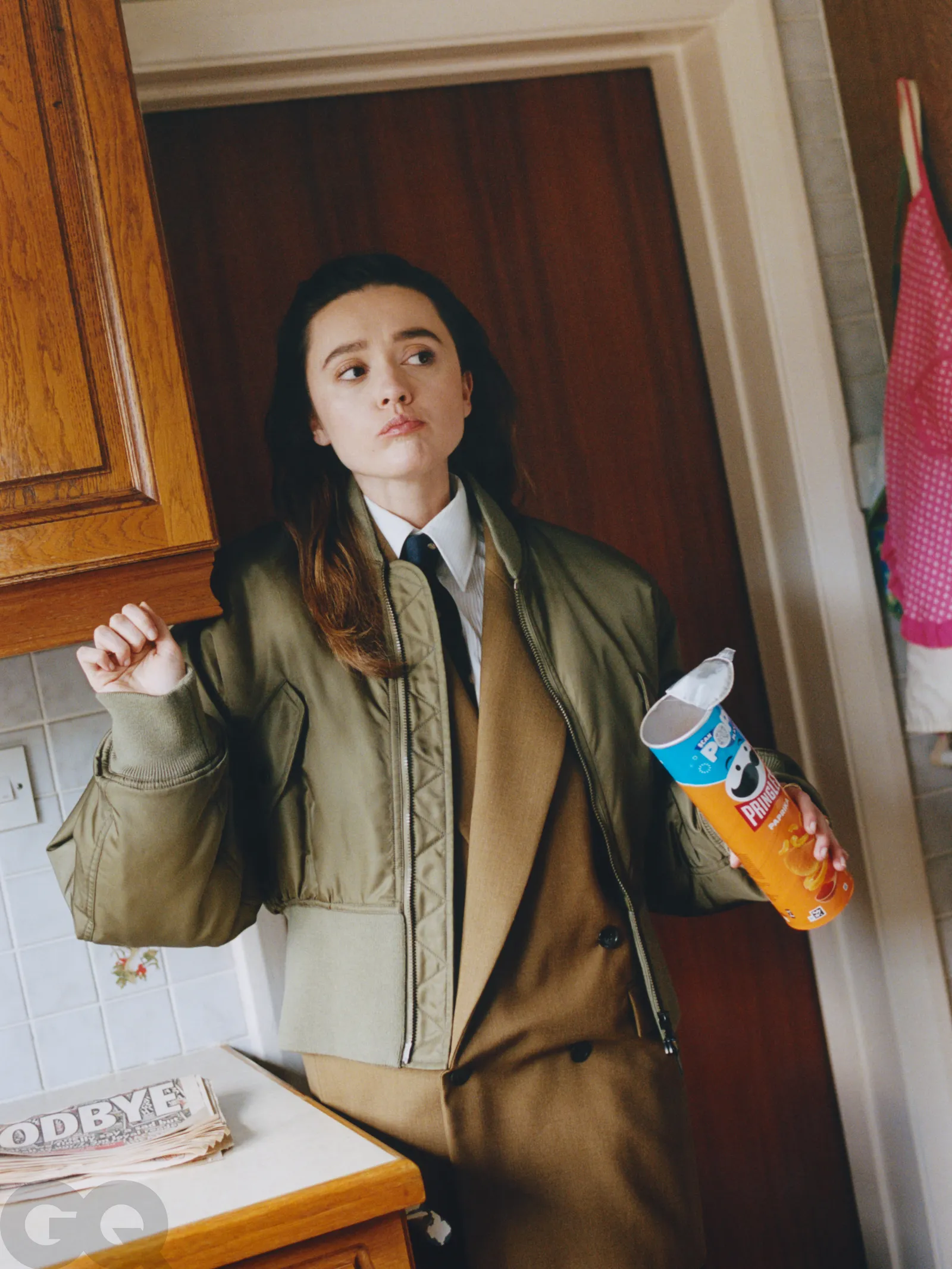 Aimee Lou Wood in a tan suit and olive bomber jacket, holding a can of Pringles in a kitchen with 90s decor.