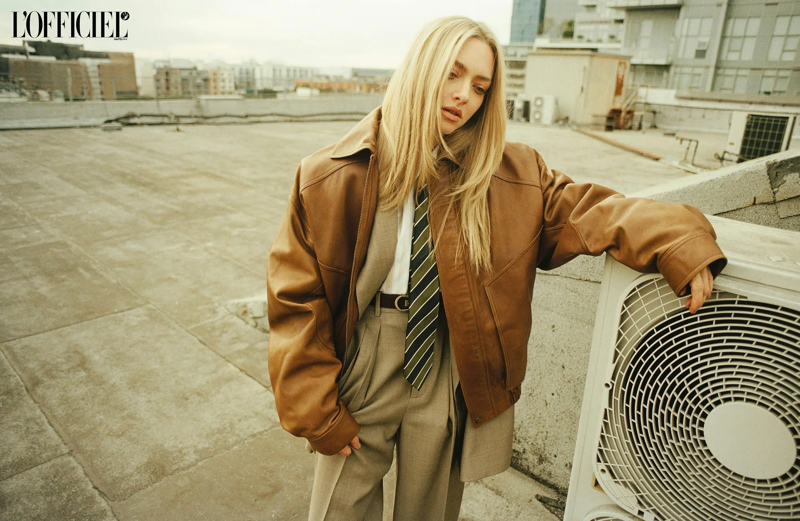 Amanda Seyfried styled in a brown leather jacket, wide-legged trousers, and striped tie on an urban rooftop scene