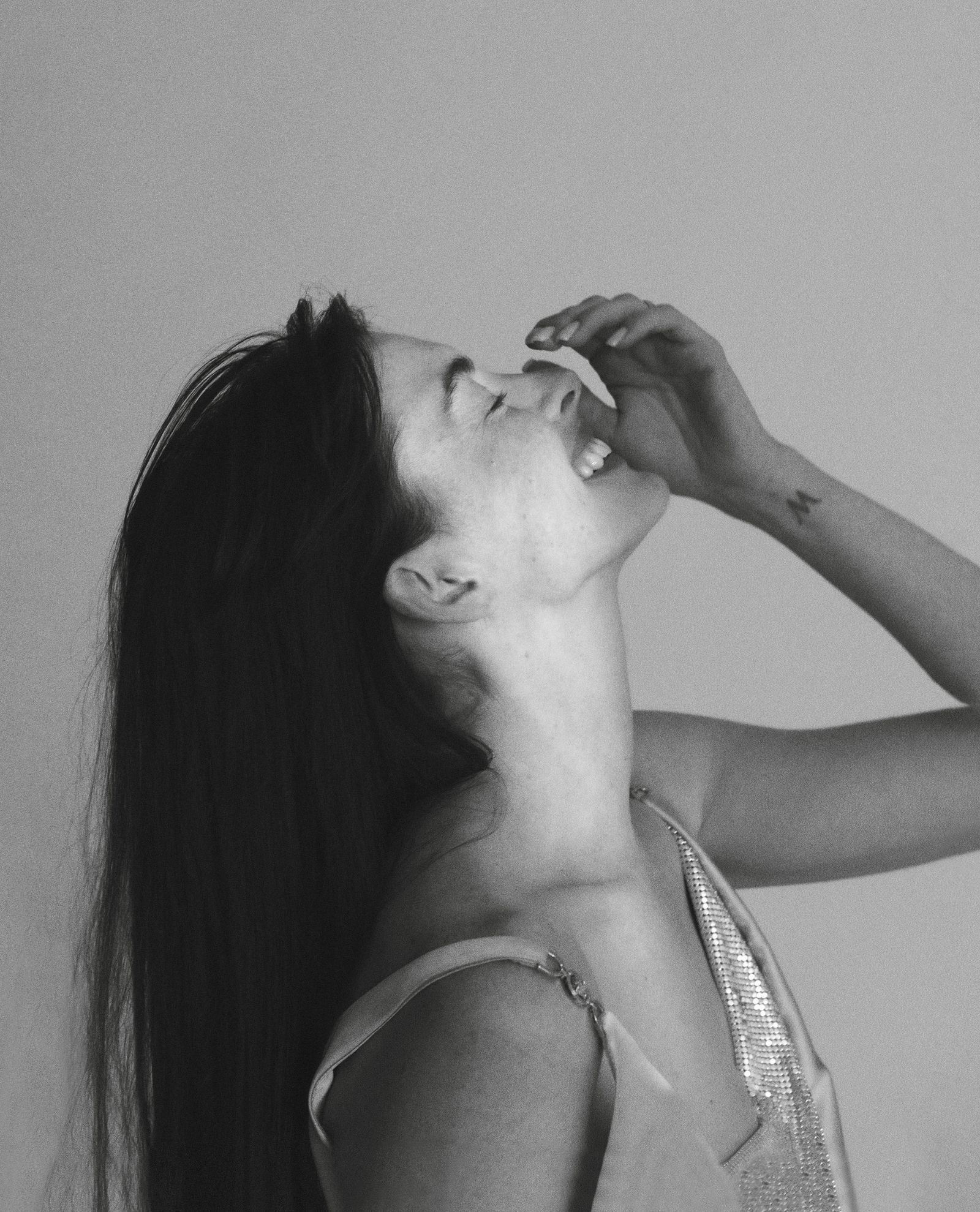 Side profile of Anne Hathaway laughing with eyes closed, wearing a shimmering silver-strap dress in a minimalist studio setting, Vogue France 2024.