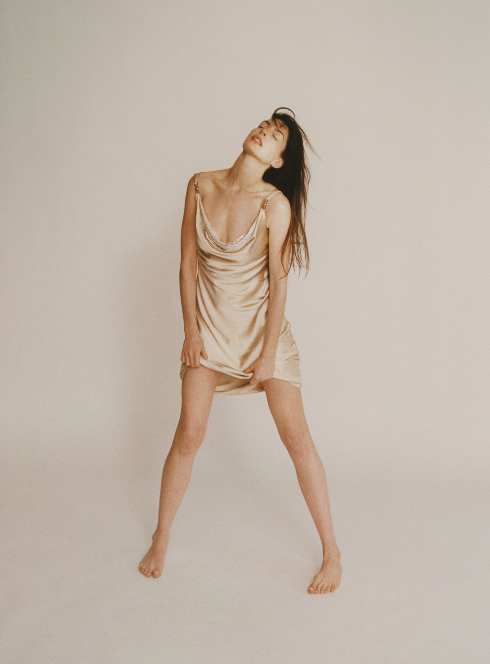 Anne Hathaway barefoot in a champagne satin mini dress, playfully tugging the hem in a neutral-toned studio shoot for Vogue France November 2024.