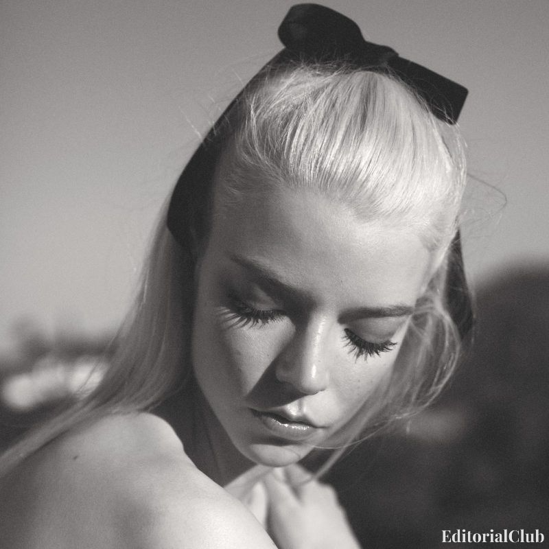 Black and white portrait of Anya Taylor-Joy with dramatic lashes and a black ribbon in her platinum blonde hair, gazing down with a soft expression.