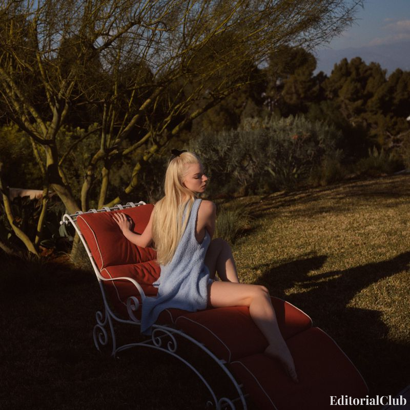 Anya Taylor-Joy sitting curled up on a red pool lounger, wearing a blue knit dress with sunset light casting warm shadows.
