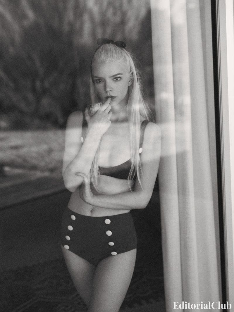 Anya Taylor-Joy in vintage-inspired high-waisted bikini posing behind glass, holding a drink and wearing a black hair bow, photographed in black and white.