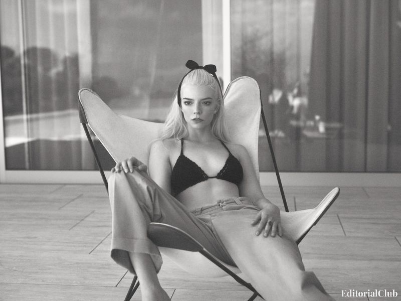 Anya Taylor-Joy sitting on a butterfly chair in a black bralette and loose trousers, styled with a bow in her hair in a monochrome editorial shot.