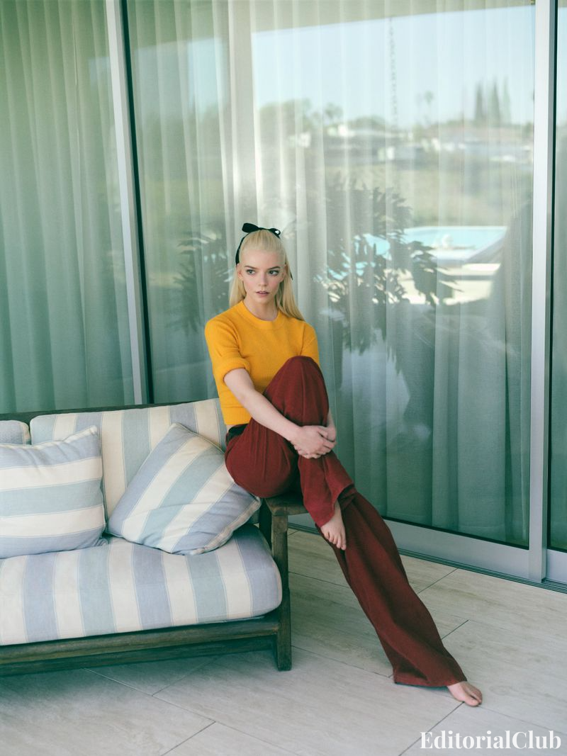 Editorial image of Anya Taylor-Joy in red trousers and a mustard yellow knit top, barefoot and seated on a striped sofa in front of sheer curtains.