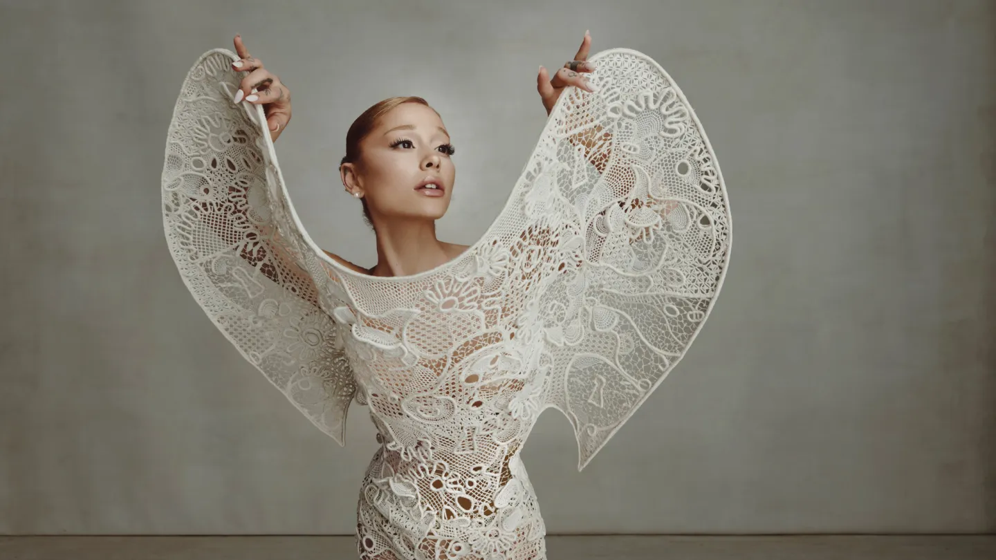 Ariana Grande posing with arms raised in a sculptural white lace gown for The Hollywood Reporter, February 2025.