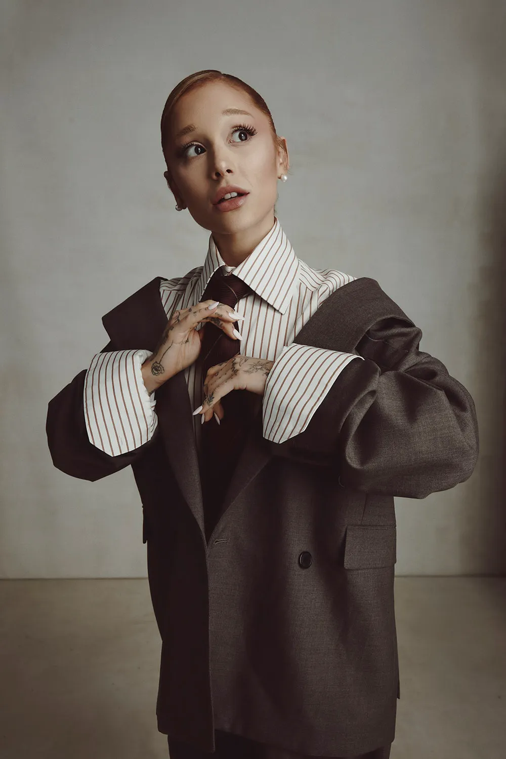 Ariana Grande wears an oversized brown suit with a striped shirt and tie, posing with expressive hands for The Hollywood Reporter February 2025.