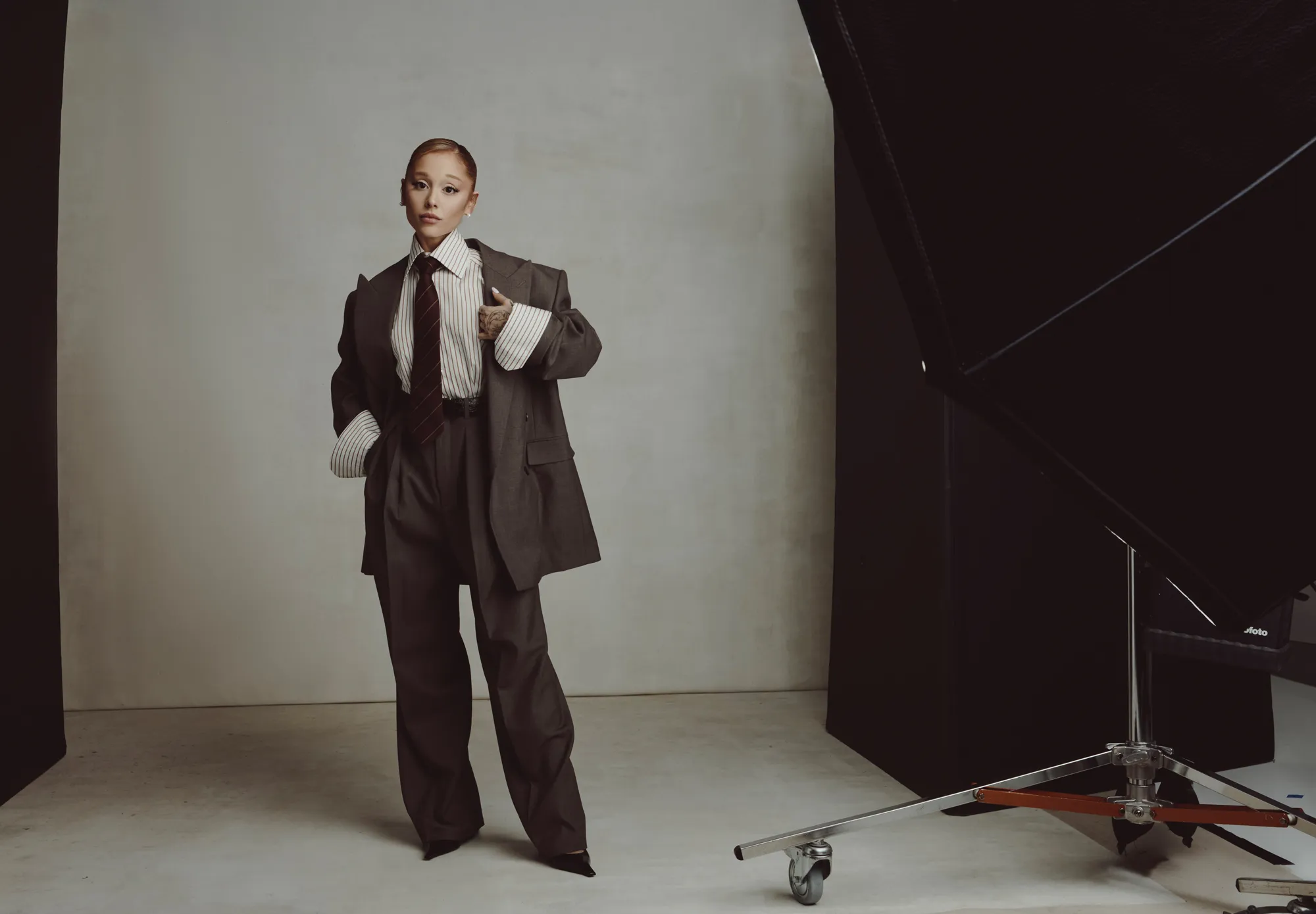 Ariana Grande in oversized pinstripe suit with wide-leg trousers and necktie in minimalist studio backdrop.
