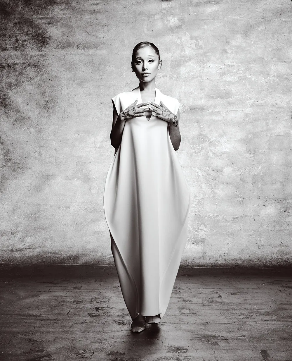 Ariana Grande standing in a minimalist set wearing a structured, sleeveless gown with hands joined, black and white portrait.
